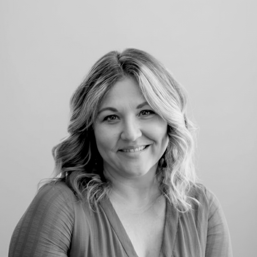 A black and white portrait of a smiling woman with wavy, shoulder-length hair, wearing a light-colored blouse, against a plain background.