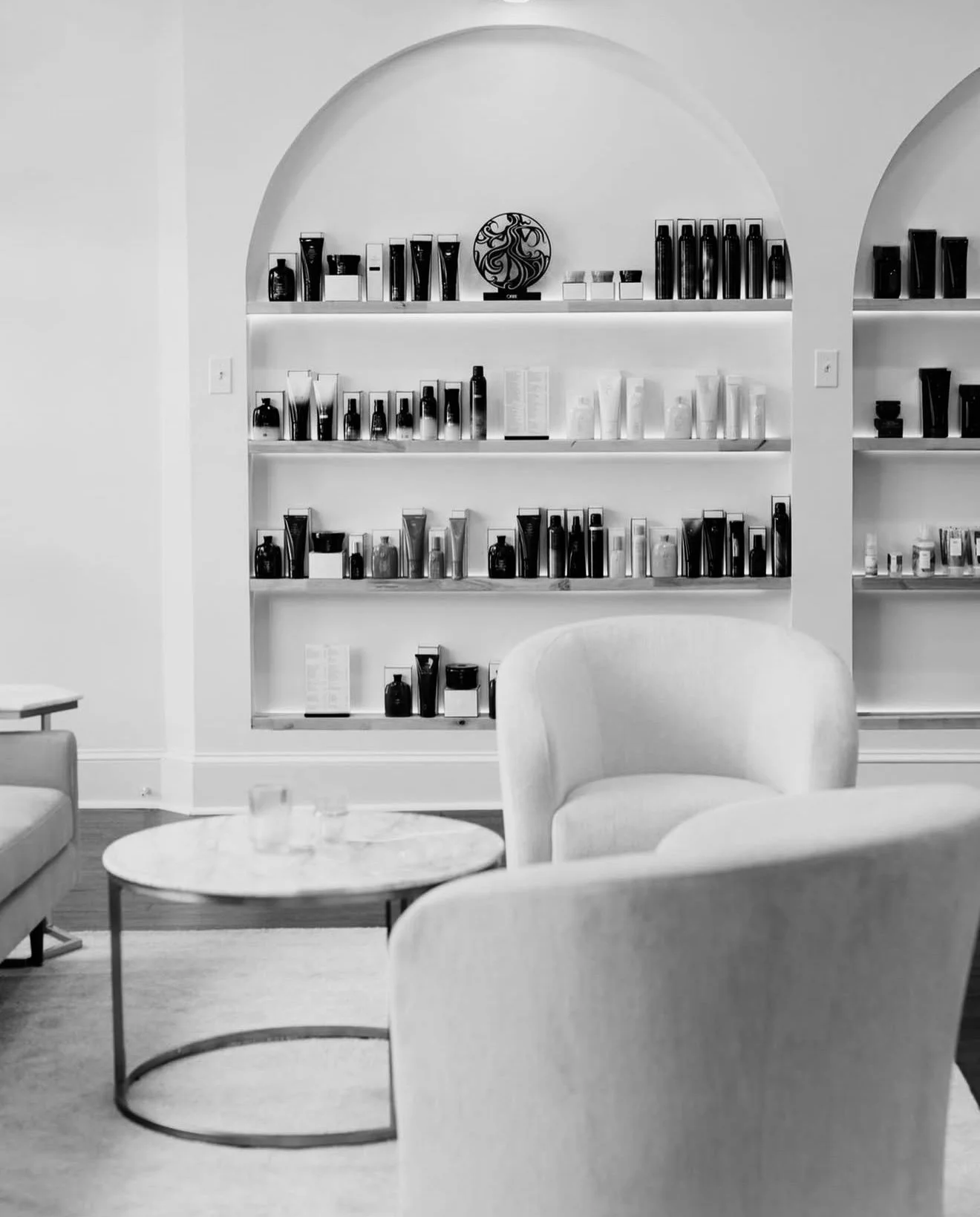 A contemporary interior with shelves filled with skincare or cosmetic products, two armchairs, and a round coffee table in black and white.