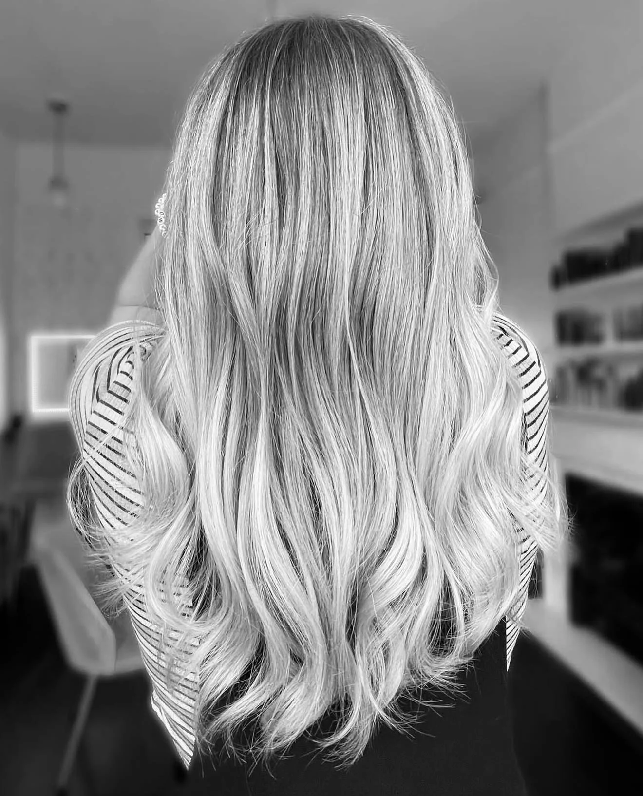 Back view of long, wavy blonde hair in a black and white photo, with a striped shirt and a blurred interior background.