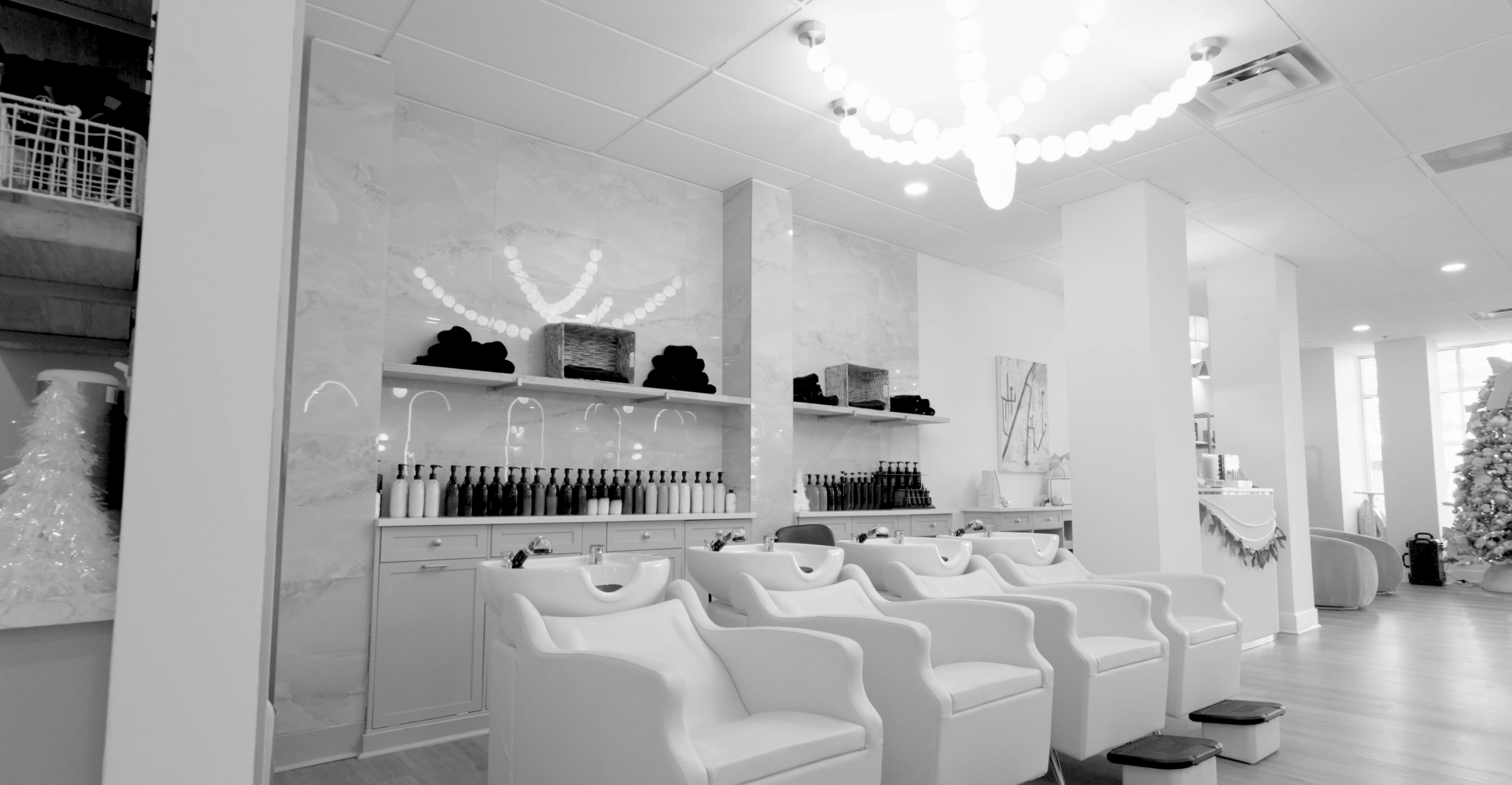 A salon with multiple white shampoo chairs and sinks, shelves with bottles, and a decorated Christmas tree in the background.