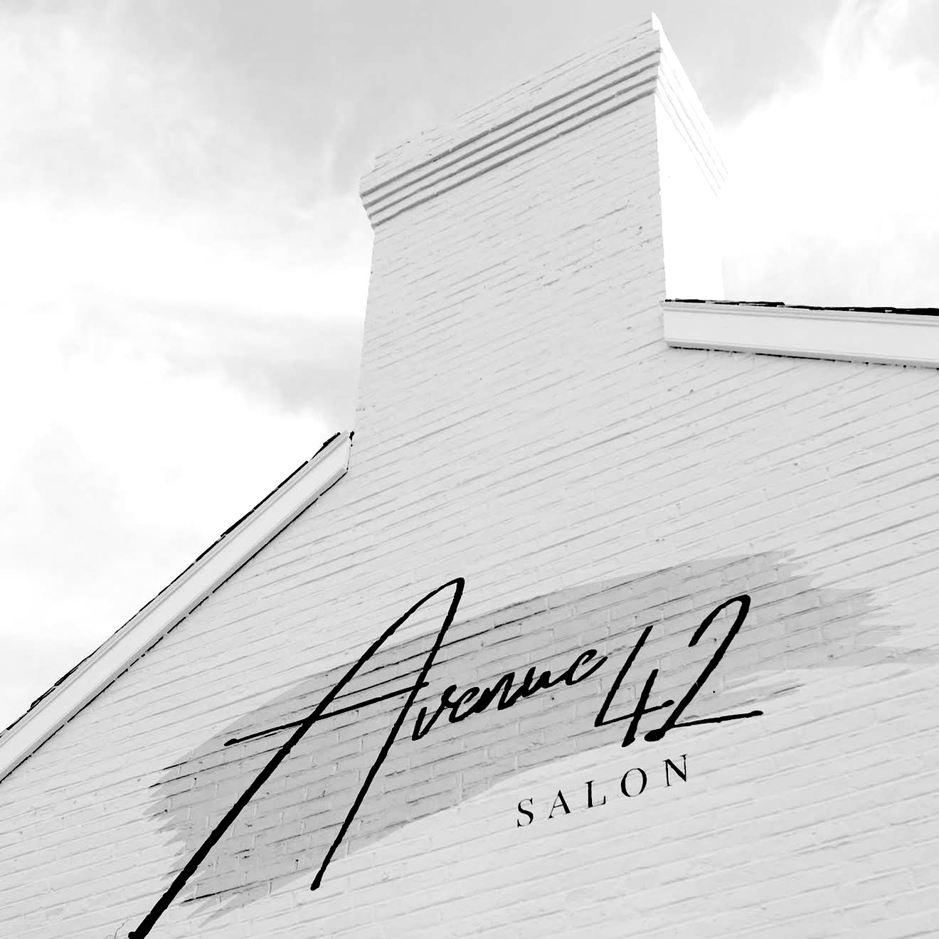 Close-up of a white brick building with a black cursive sign that reads 'Avenue 42 Salon' on the wall.