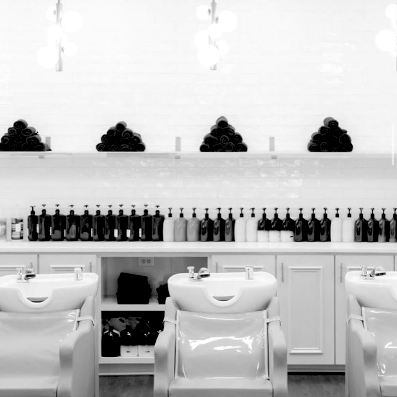Salon or barbershop interior with shampoo sinks in the foreground, shelves with hair care products above, and stacks of towels and bottles on the wall.