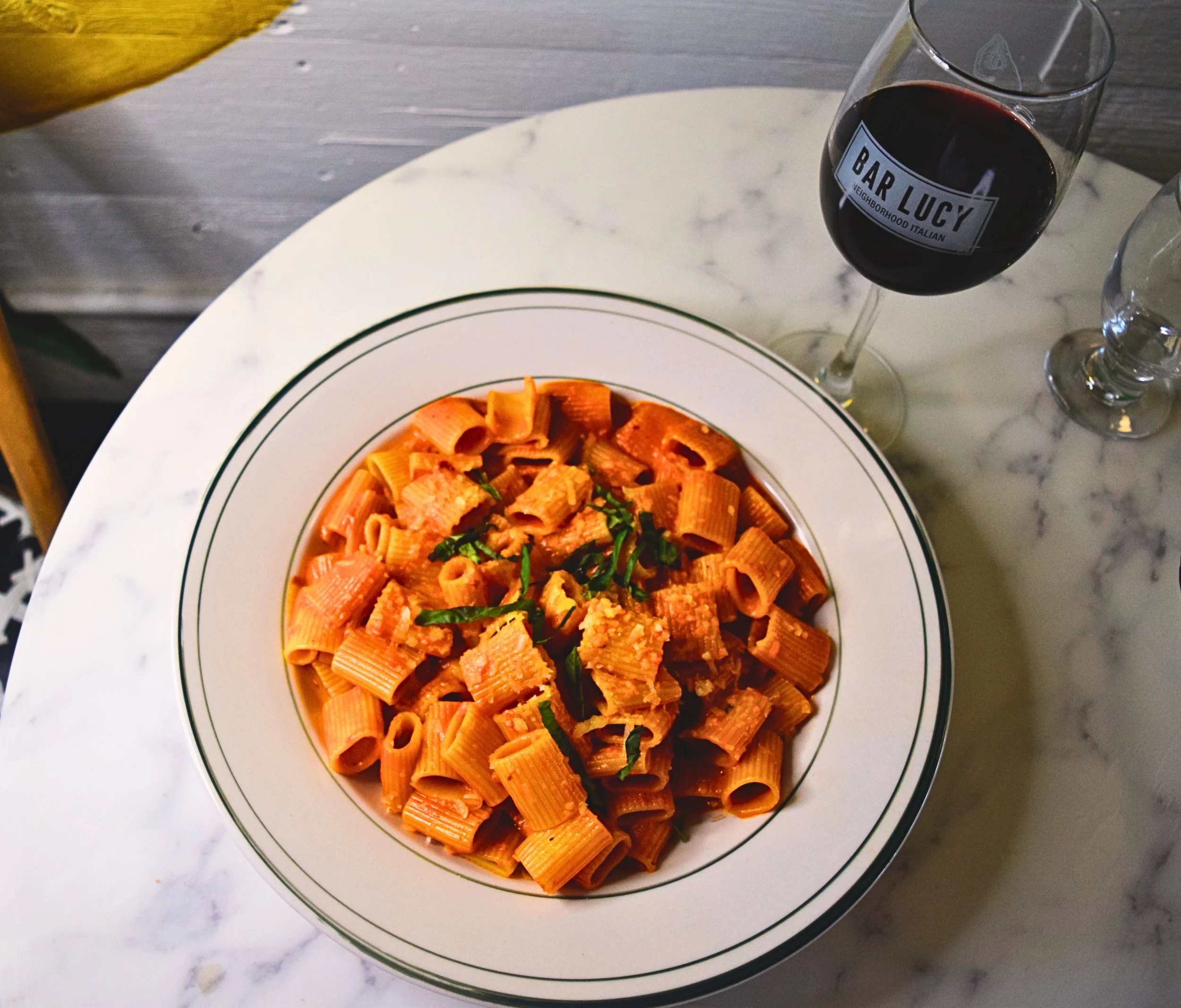 Plate of rigatoni pasta with tomato sauce and basil, a glass of red wine, and a glass of water on a white marble table.