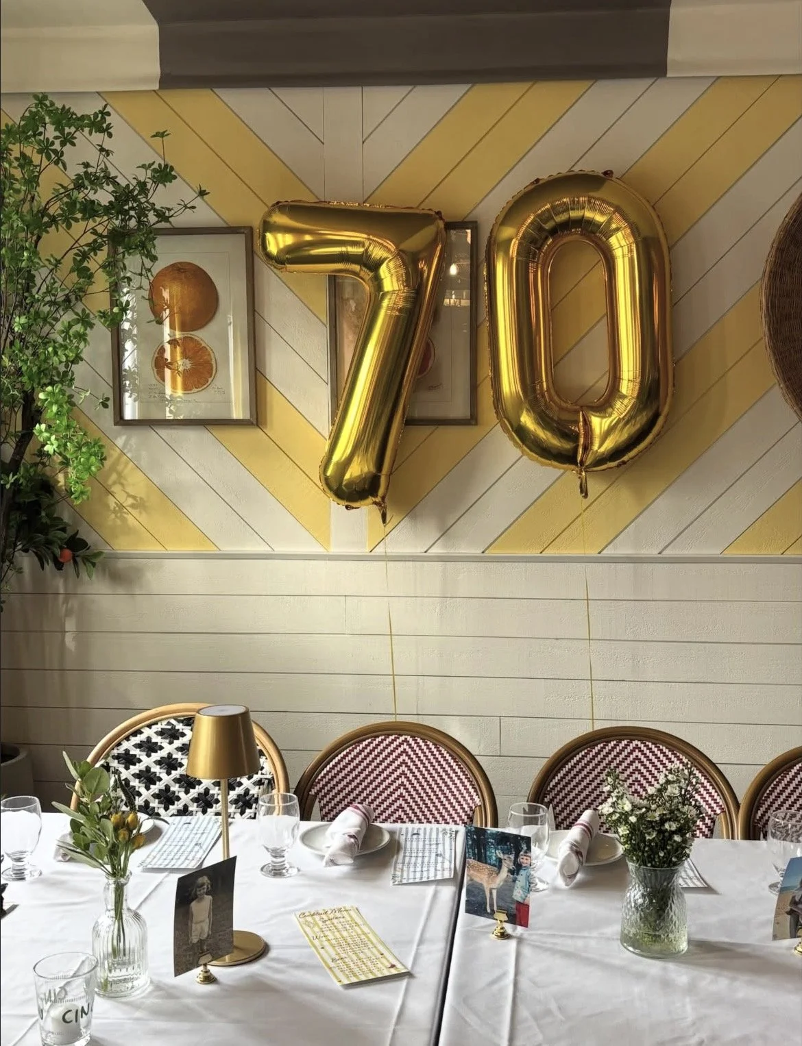 Gold balloons forming the number 70 hang on a decorated wall with plants and framed artwork. A dining table set with white tablecloth, floral arrangements, glasses, and placemats sits in front of the wall.