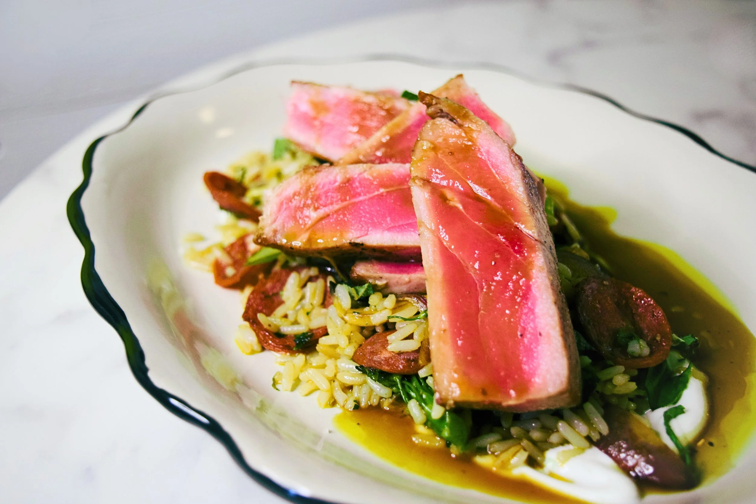 A plate of grilled tuna steak served over seasoned rice with vegetables and a drizzle of sauce.