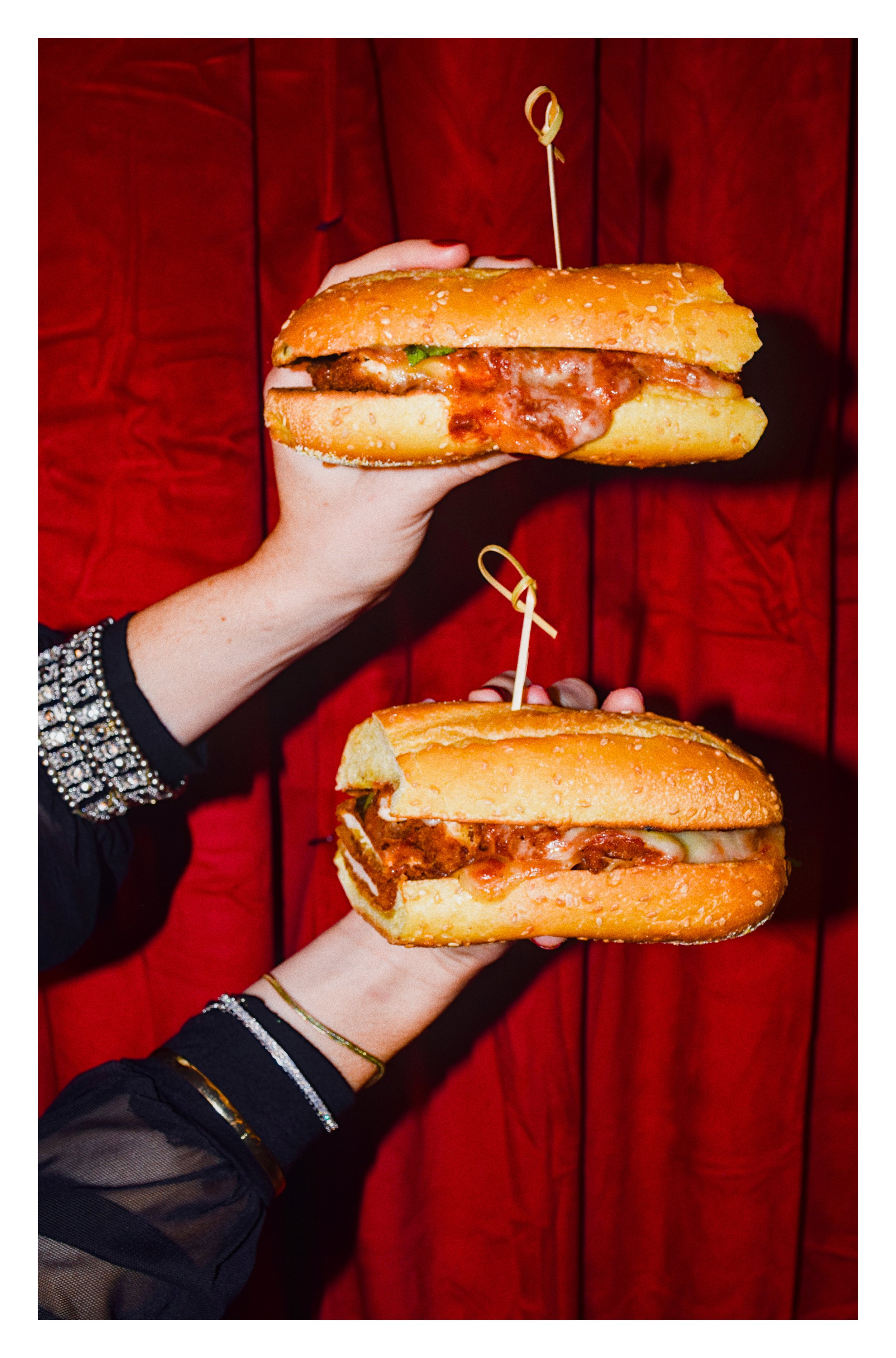 Two hands each holding a large sandwich with sesame seed bun, cheese, lettuce, and meat, against a red wooden background.