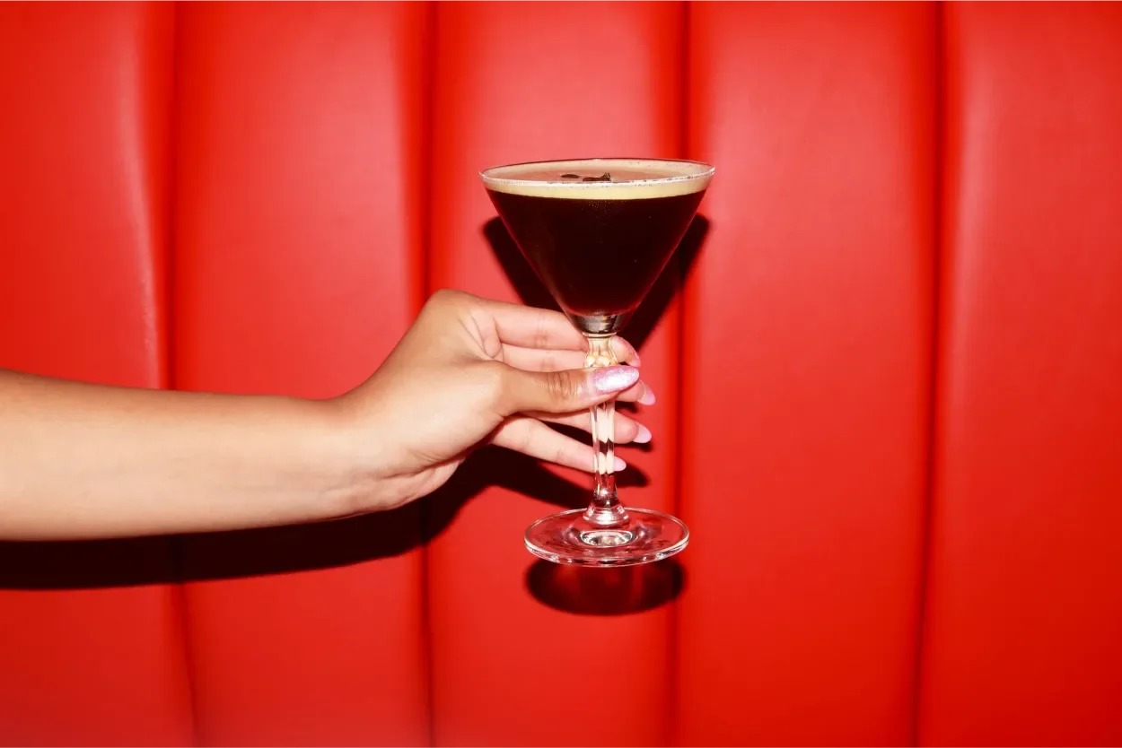 A person's hand holding a cocktail glass with a dark beverage against a red upholstered background.