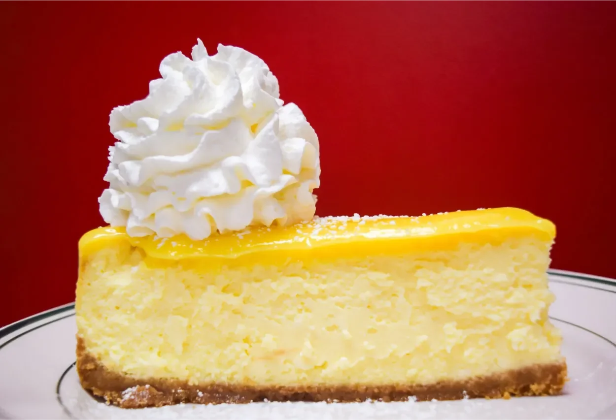 A slice of lemon cheesecake with whipped cream on top, sitting on a white plate with a decorative border.