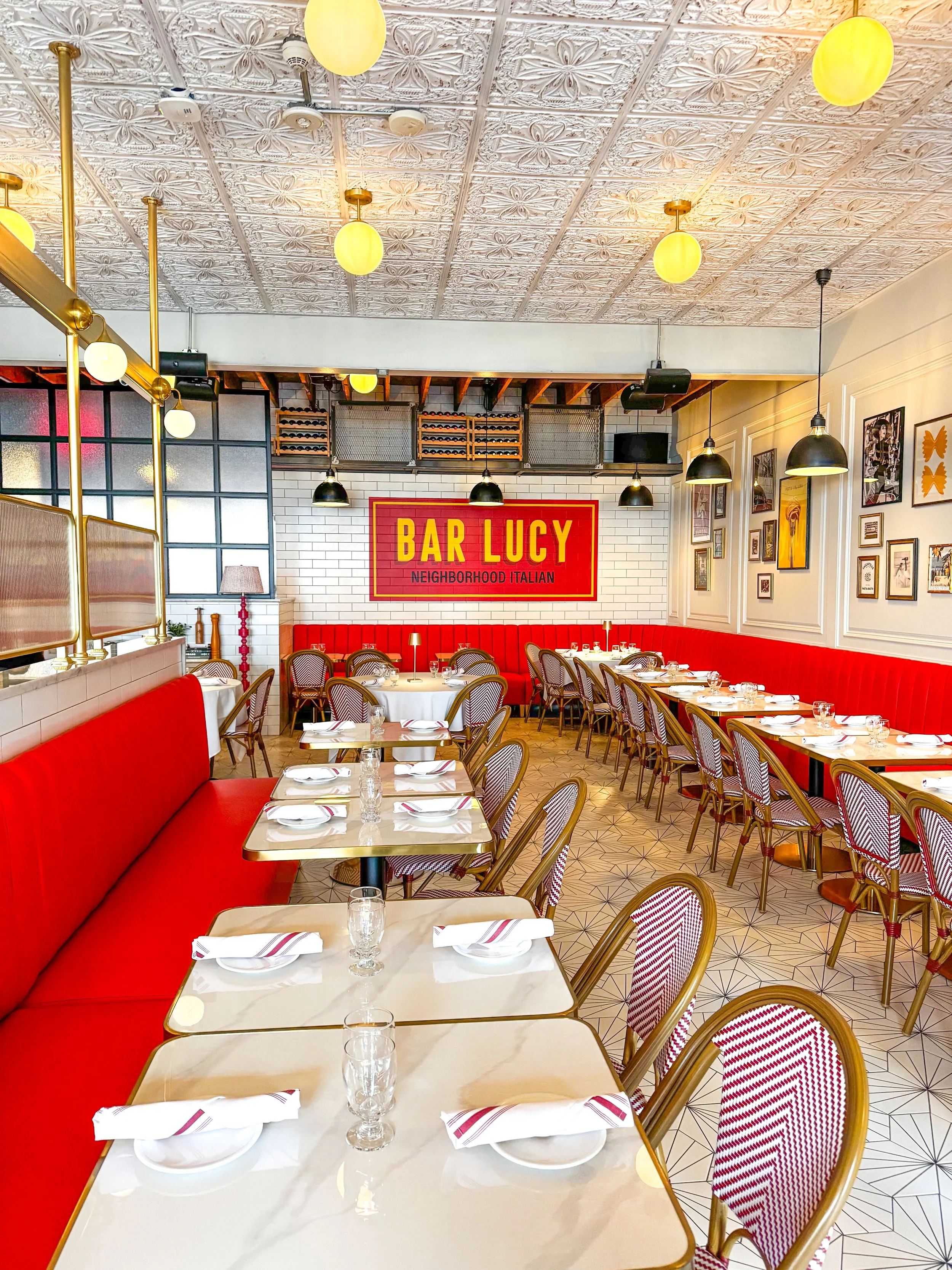 Inside Bar Lucy's dining room with red upholstered seating and tables set with plates, glasses, and napkins, featuring our hand painted logo, that reads "Bar Lucy Neighborhood Italian"