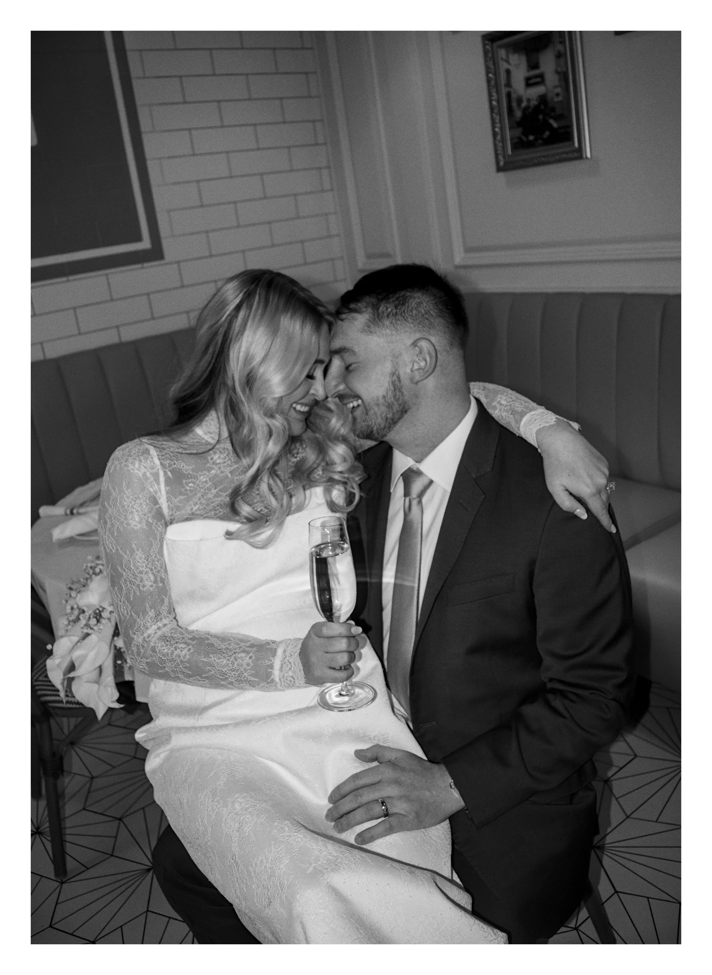 A black and white photo of a bride and groom celebrating their wedding. The bride is wearing a lace wedding dress and holding a glass of champagne. They are seated close together, touching foreheads and smiling. The bride has blonde hair in loose cur