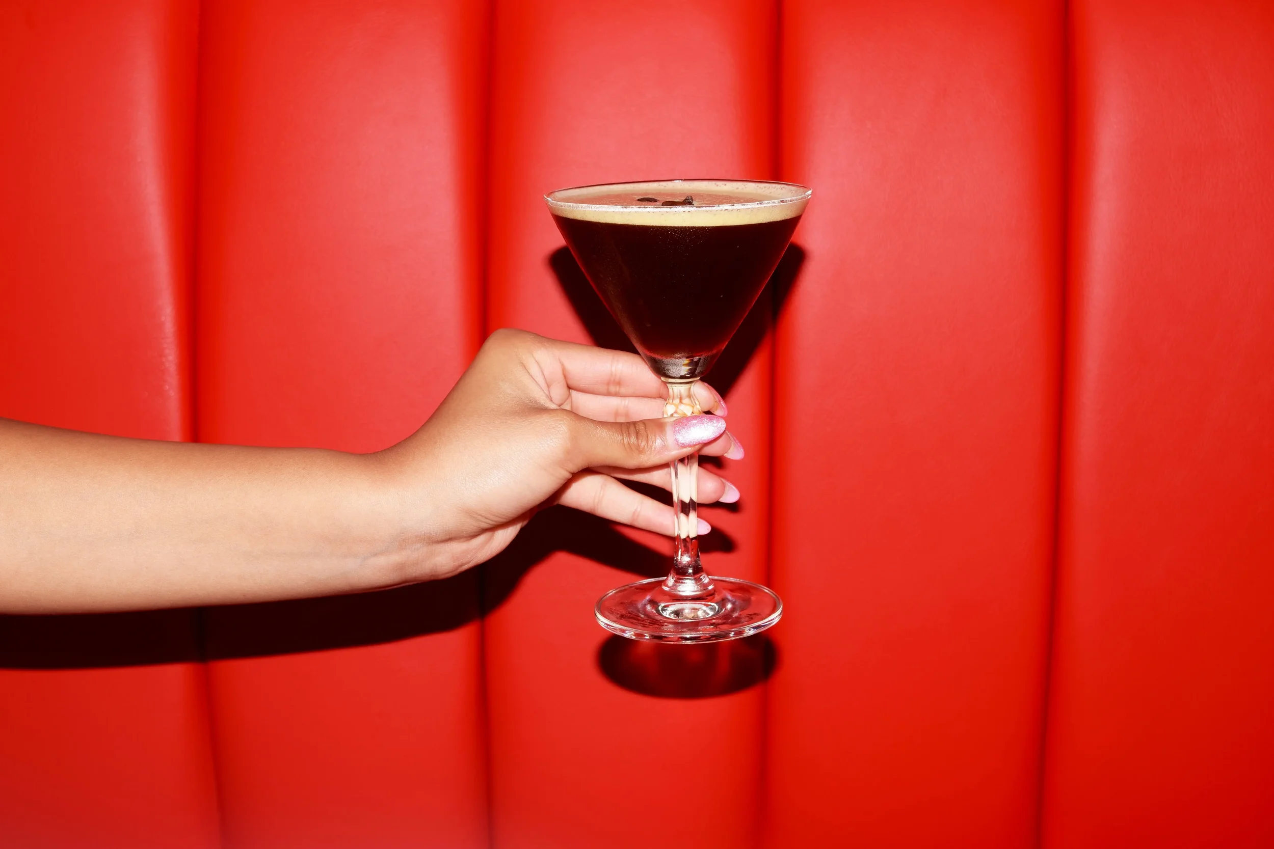 A hand holding a martini glass filled with a dark coffee-colored beverage against a red padded background.
