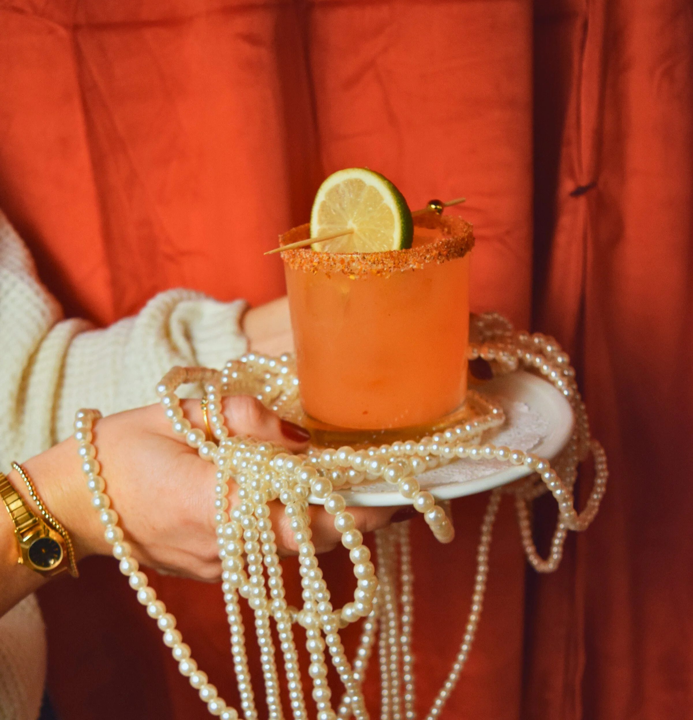 Person holding a plate with a margarita glass filled with orange drink, garnished with a lime wedge, surrounded by pearl necklaces, against a burgundy background.