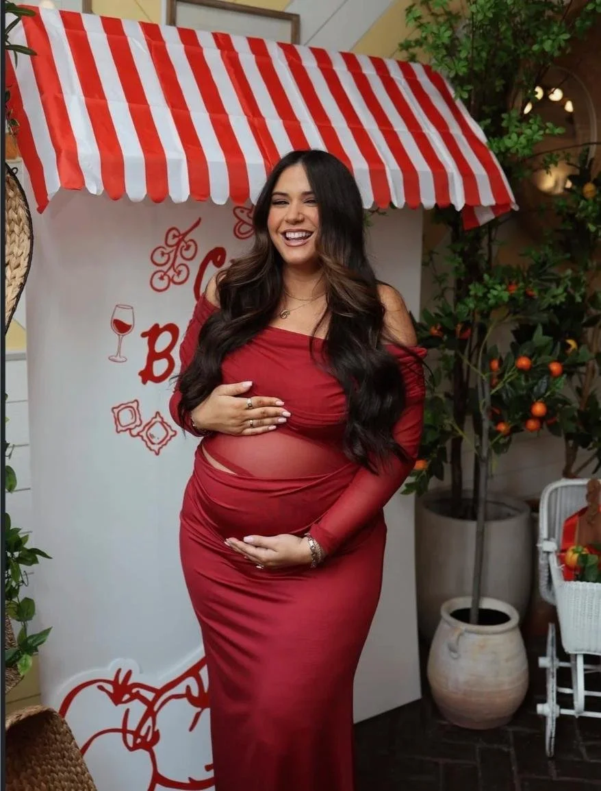 Pregnant woman in a red dress smiling at a birthday or baby shower celebration, holding her belly, standing outdoors near greenery and a decorated backdrop.