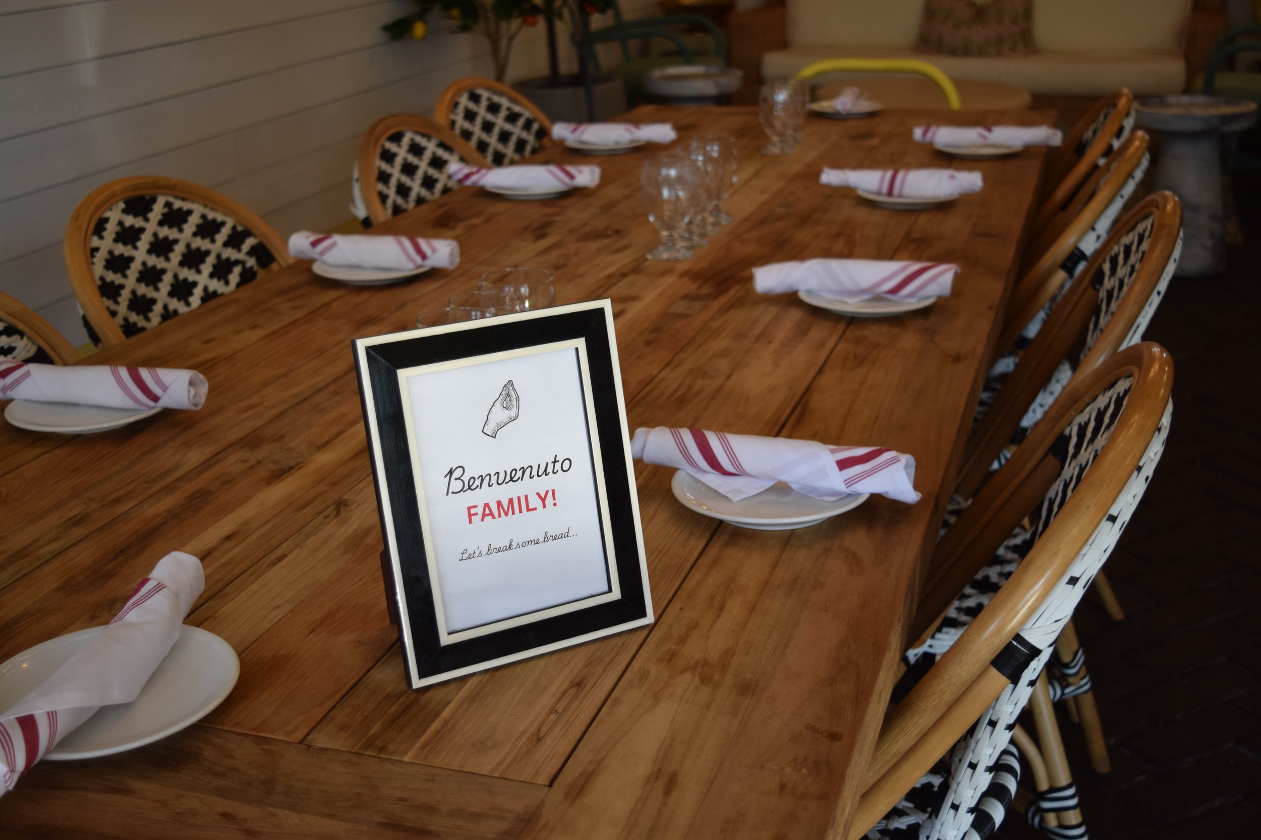 A long wooden dining table set with white plates, napkins, and glasses, with a framed sign in the foreground reading "Benvenuto FAMILY! Let's break some bread..."