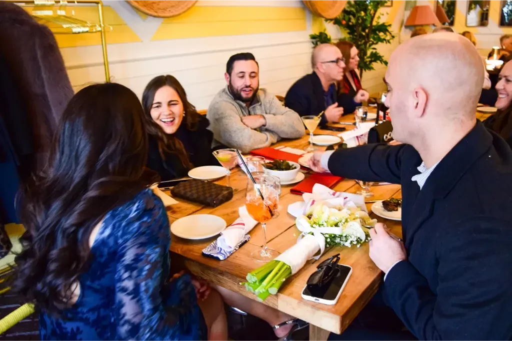 Group of people sitting around a wooden table in a restaurant, smiling and enjoying drinks, with a floral bouquet, plates, and napkins on the table.