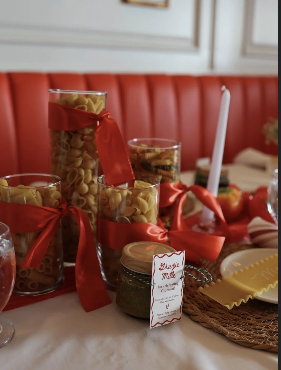Pasta in glass jars with red ribbons, a jar of herbs, and a white candle on a decorated table for a celebration.