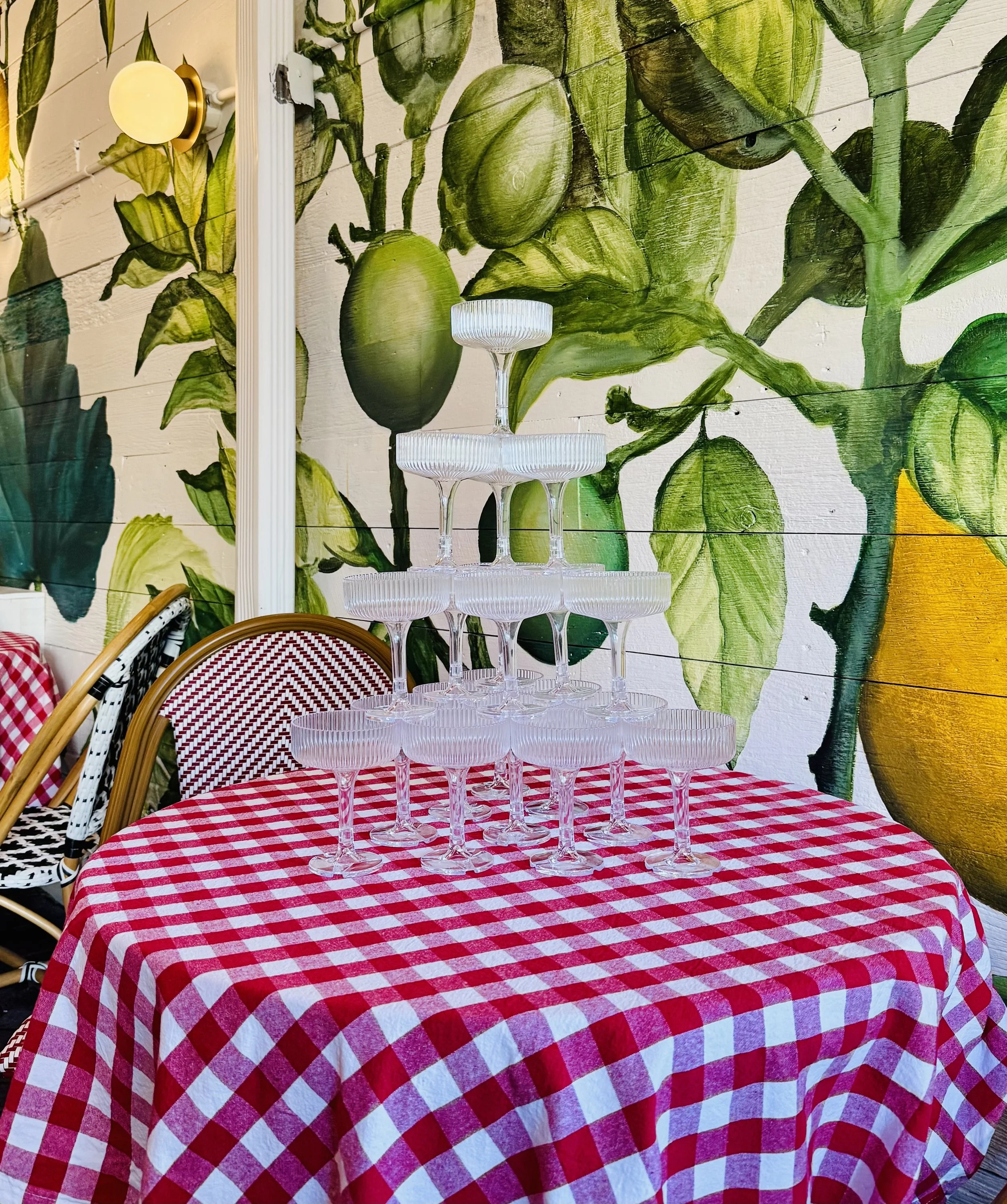 Stacked glasses arranged in a pyramid on a red and white checkered tablecloth with a colorful leaf and lemon mural in the background.