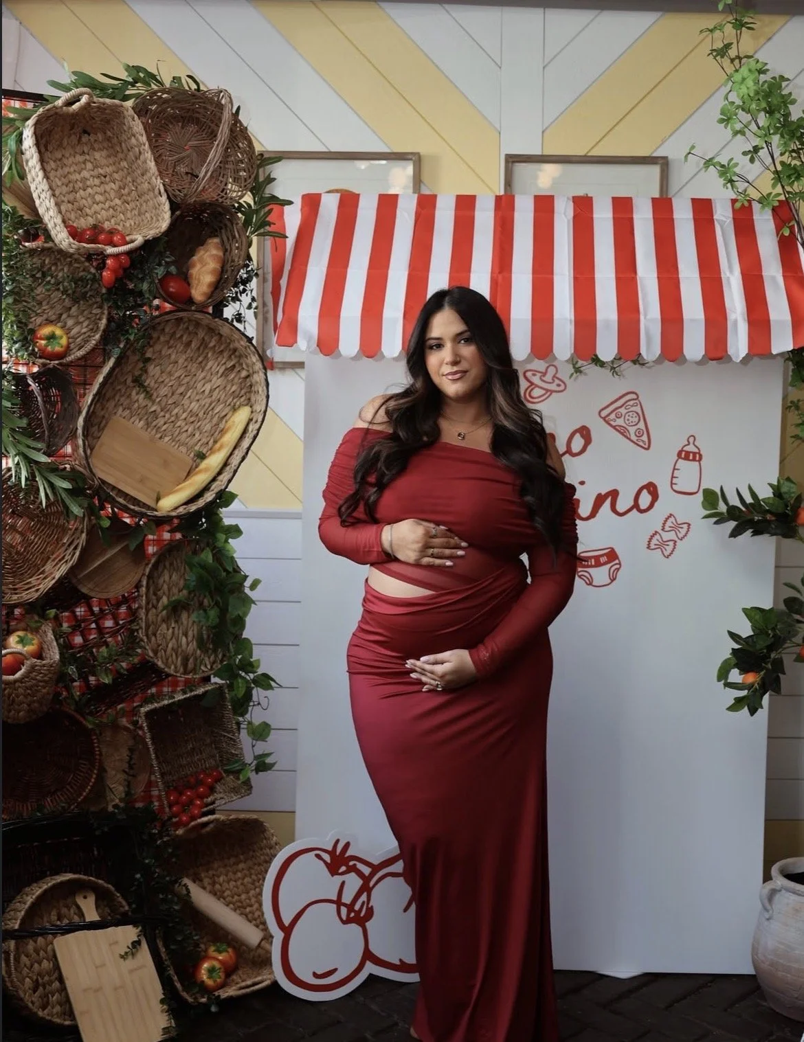 A woman standing in front of a red and white striped canopy at a food or farmers market, with woven baskets of produce on the left side and a white backdrop with red writing and illustrations behind her.