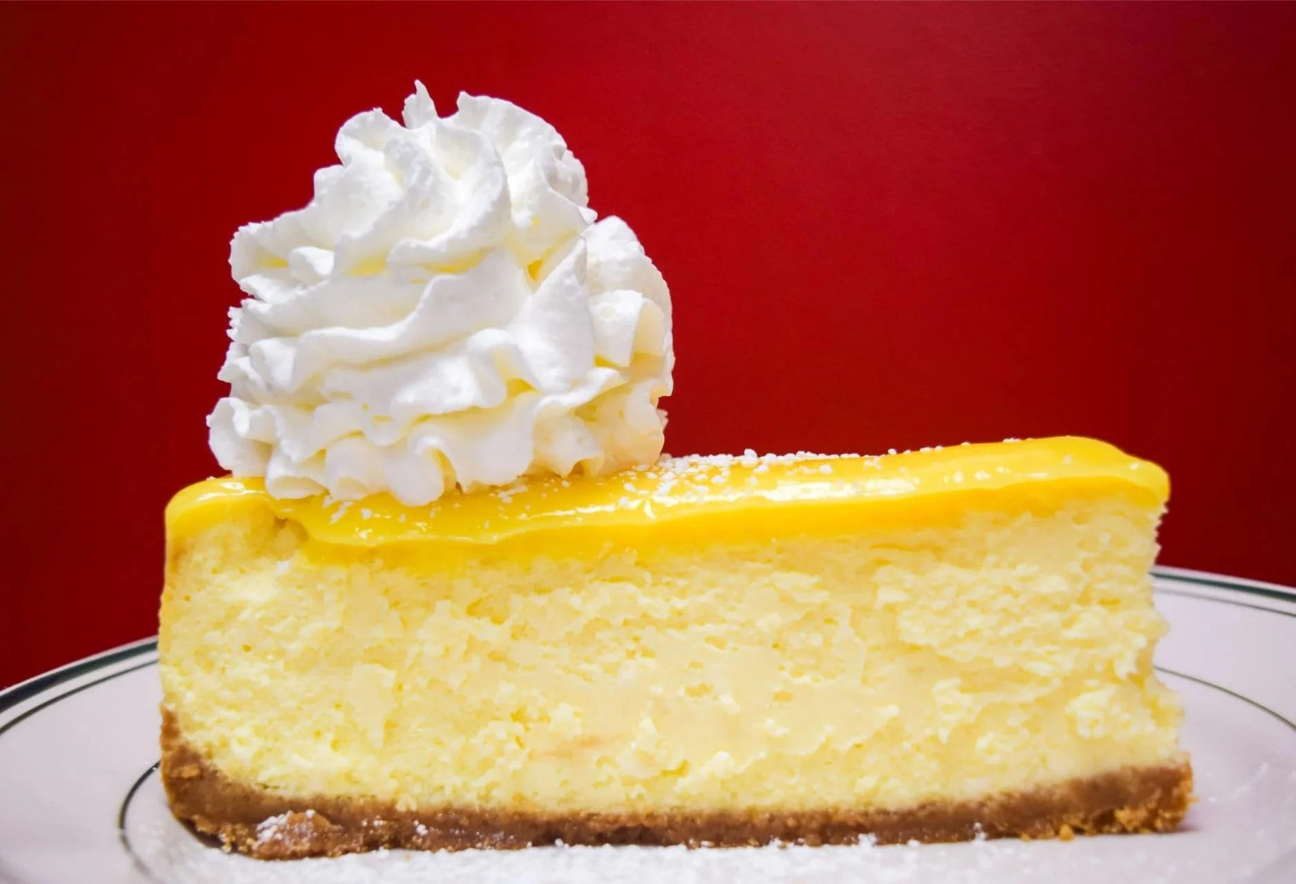 A slice of lemon cheesecake topped with whipped cream on a white plate with a red background.