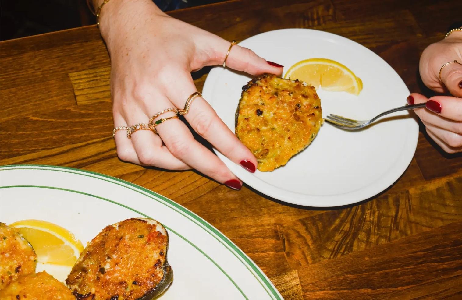 Person holding a white plate with a baked stuffed mushroom cap and a lemon wedge, with another plate of similar dish on a wooden table.
