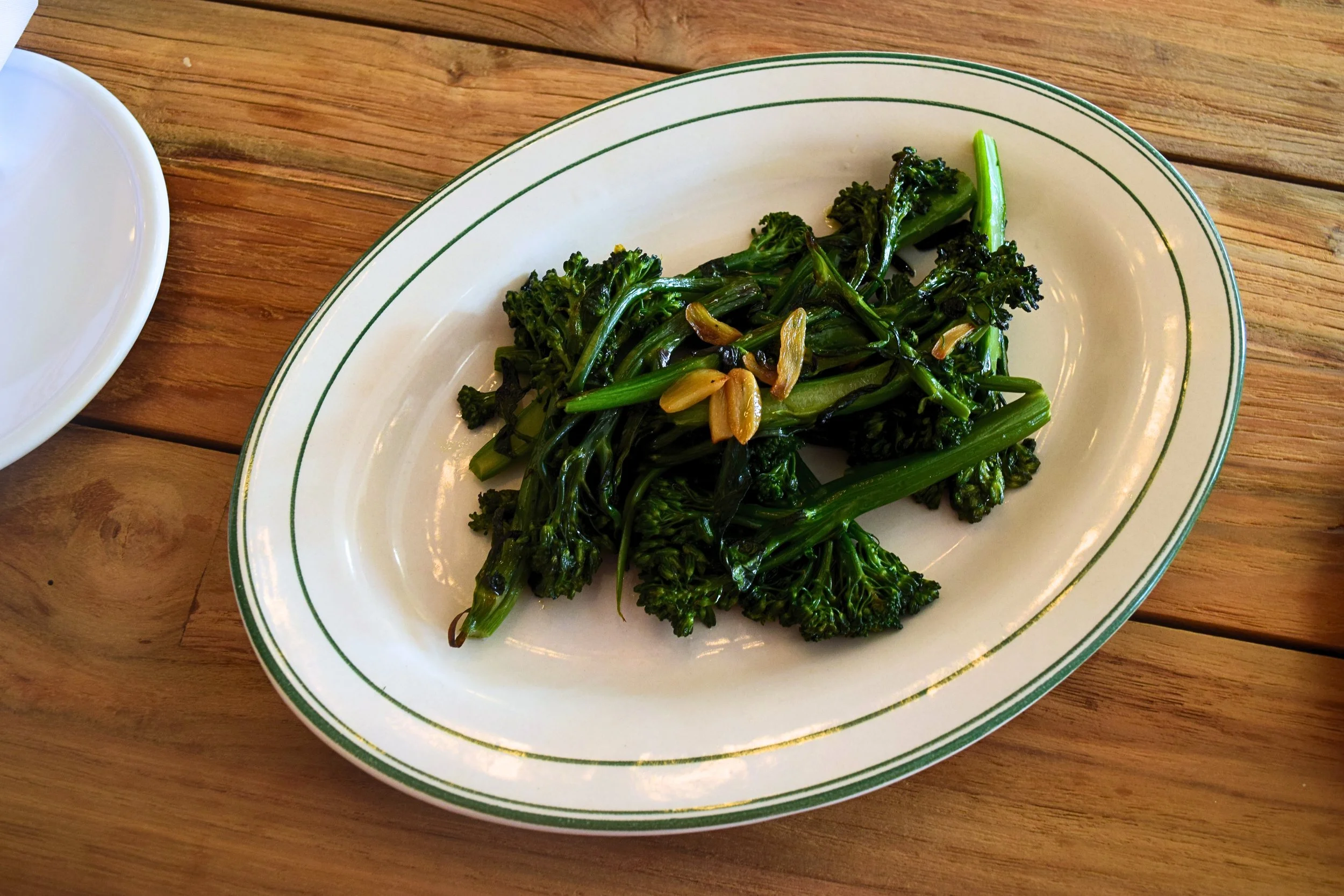 Cooked broccolini with sliced garlic on a white oval plate with green trim, placed on a wooden table.