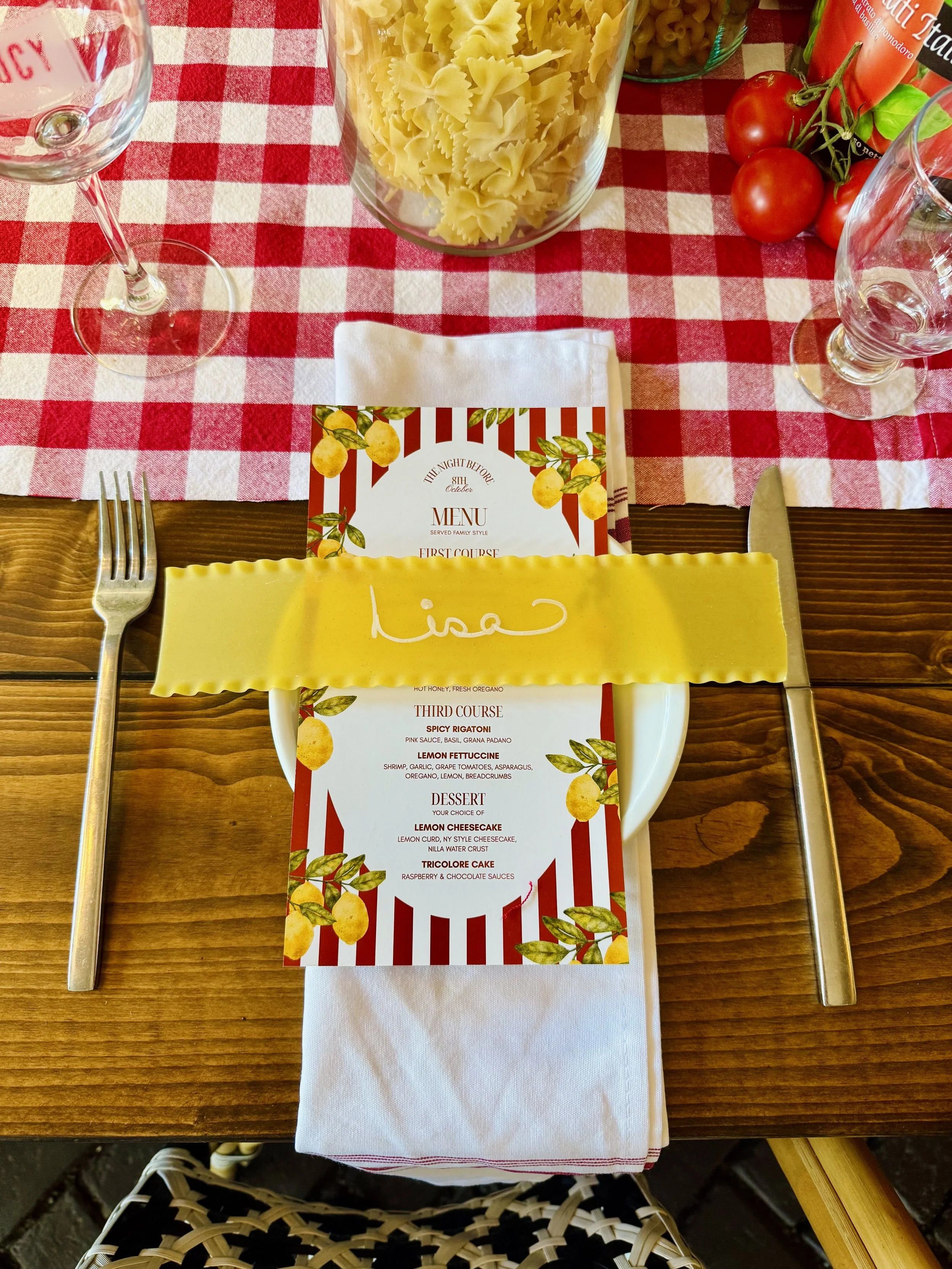 A table setting with a striped menu on a white plate, a yellow pasta-like napkin with a handwritten name, a fork and knife, a wine glass, a glass jar of pasta, cherry tomatoes, and a checkered tablecloth.