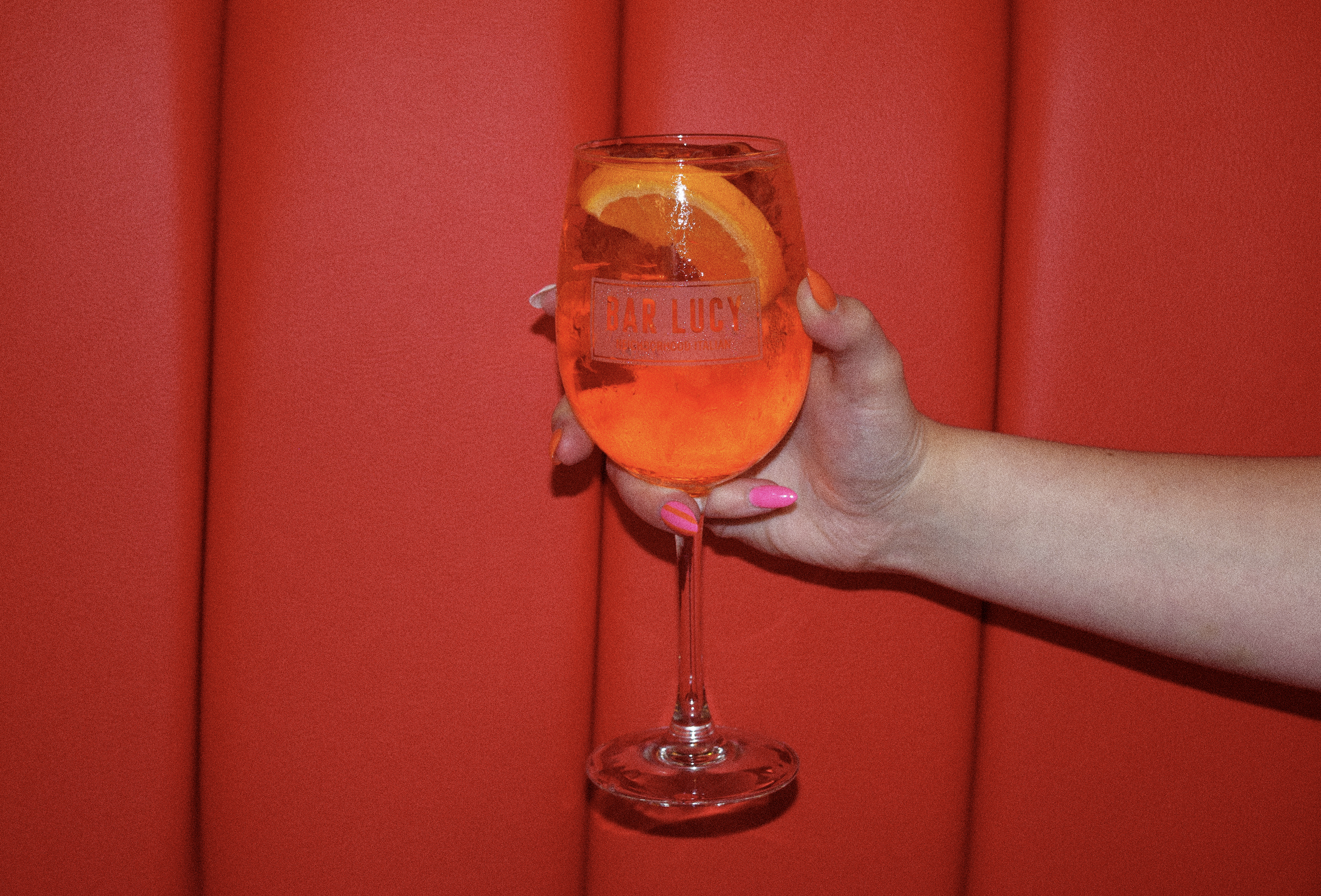 Hand holding a wine glass filled with an orange-colored beverage, garnished with orange slices, against a red background.