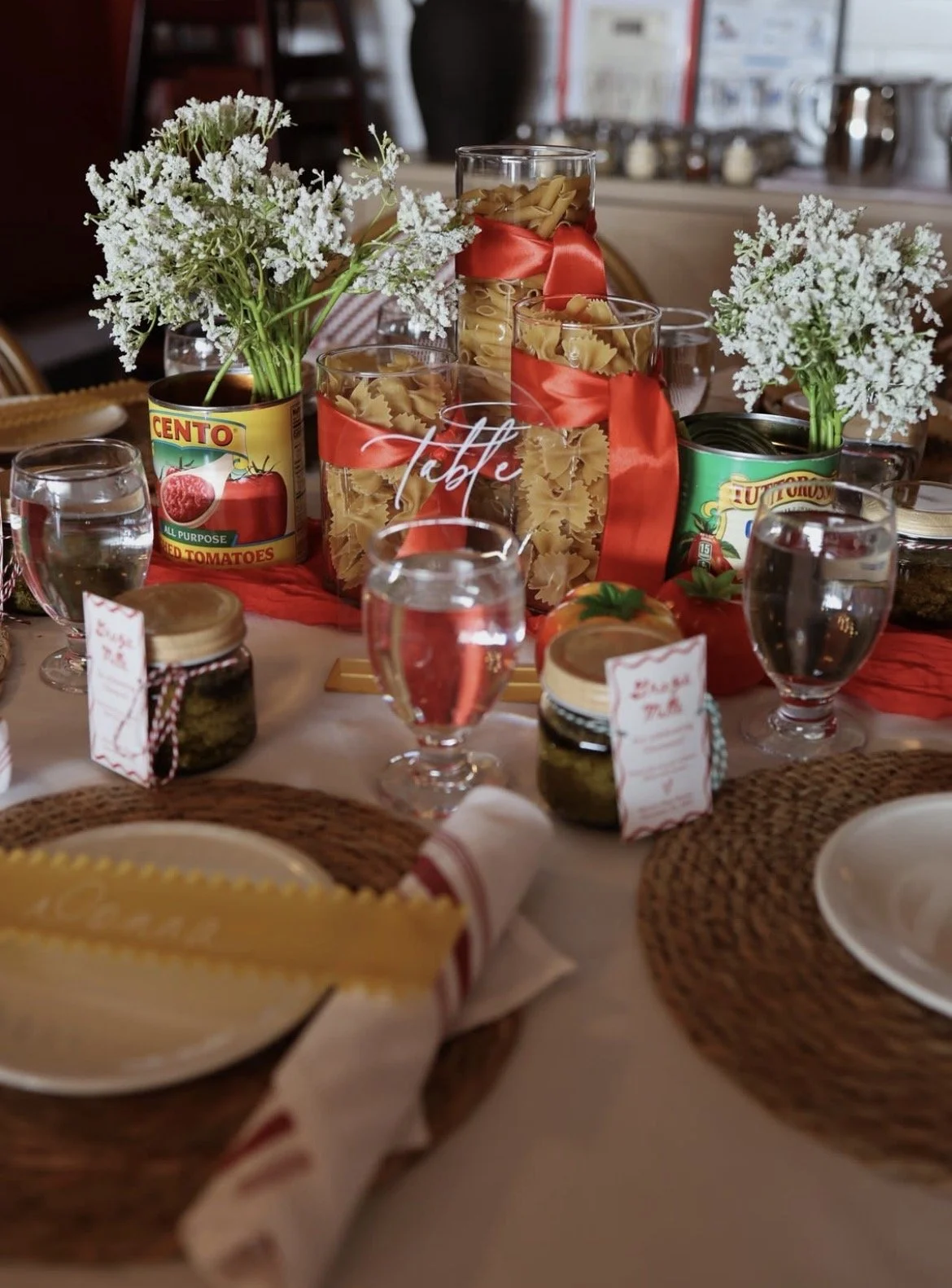 Table setting with pasta jars, flowers, water glasses, and a decorative centerpiece, ready for a meal.