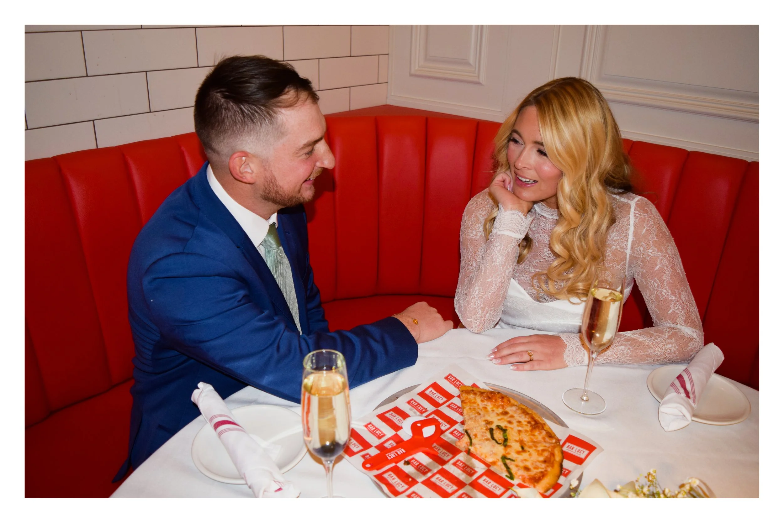 A man and woman sitting at a restaurant table with a slice of pizza and two glasses of champagne between them, smiling at each other.