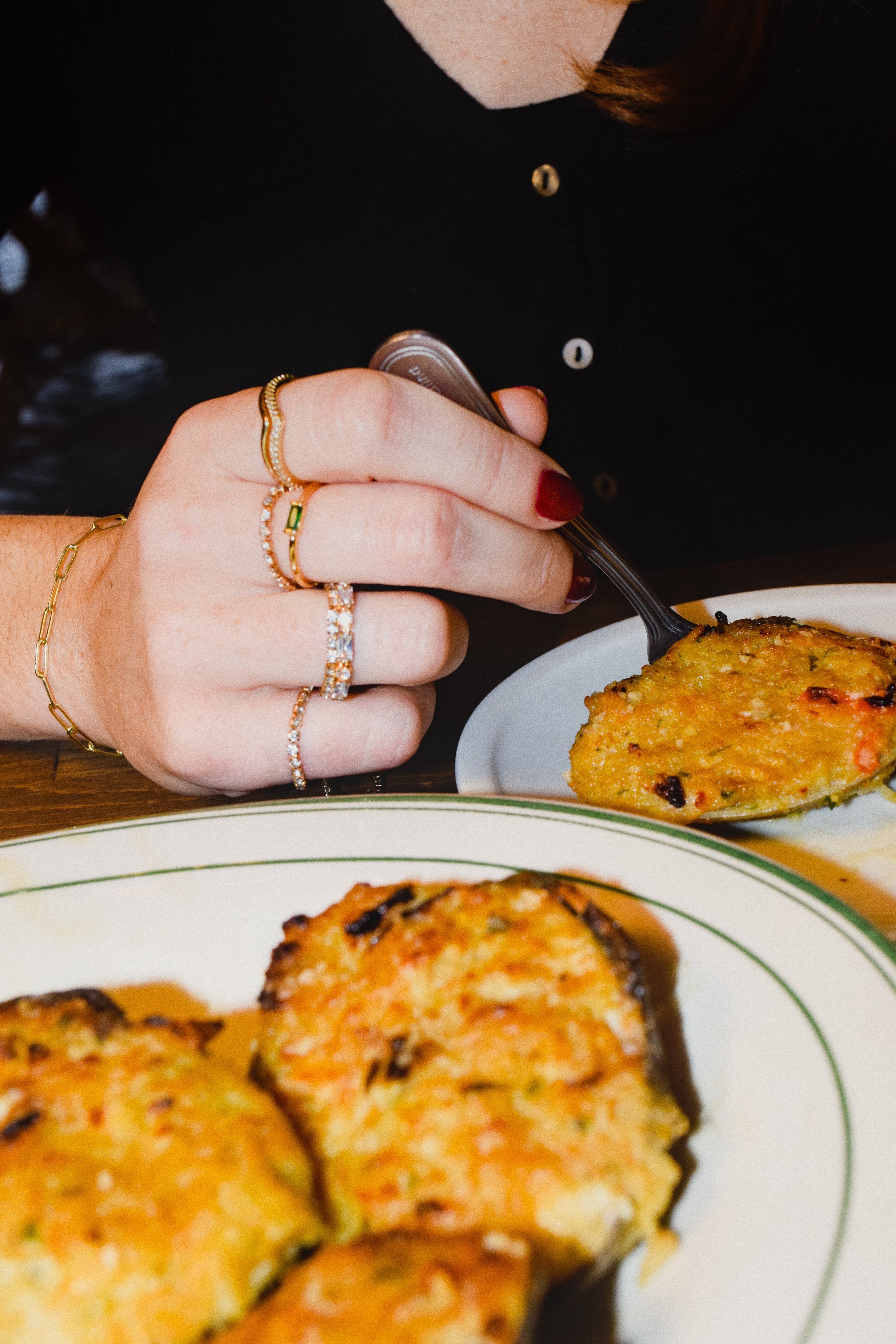 A person with red nail polish and multiple rings on their fingers is holding a spoon with a serving of baked casserole or similar dish. There are plates of the same dish on the table.