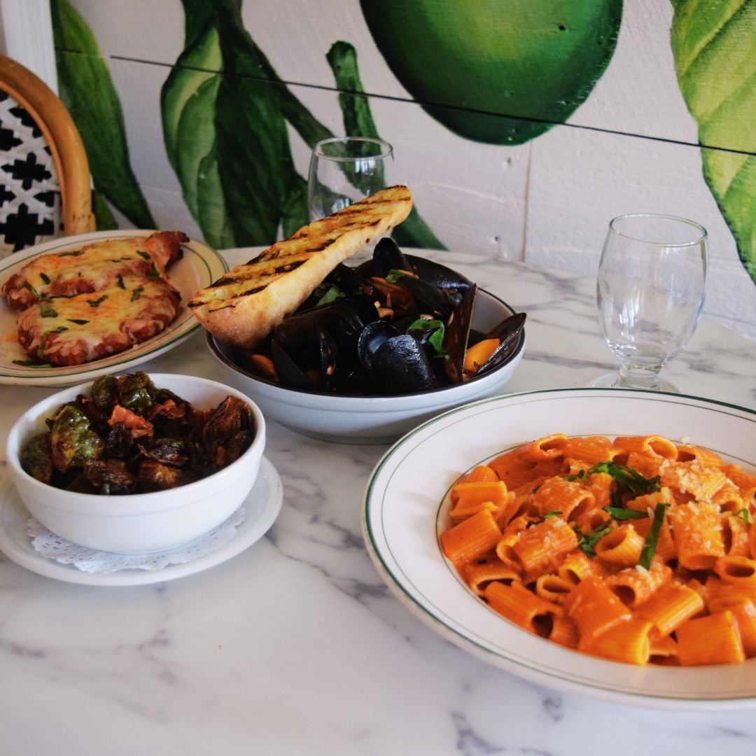 A table with Italian dishes including pasta, pizza, and seafood, along with empty glasses and a green leafy mural in the background.
