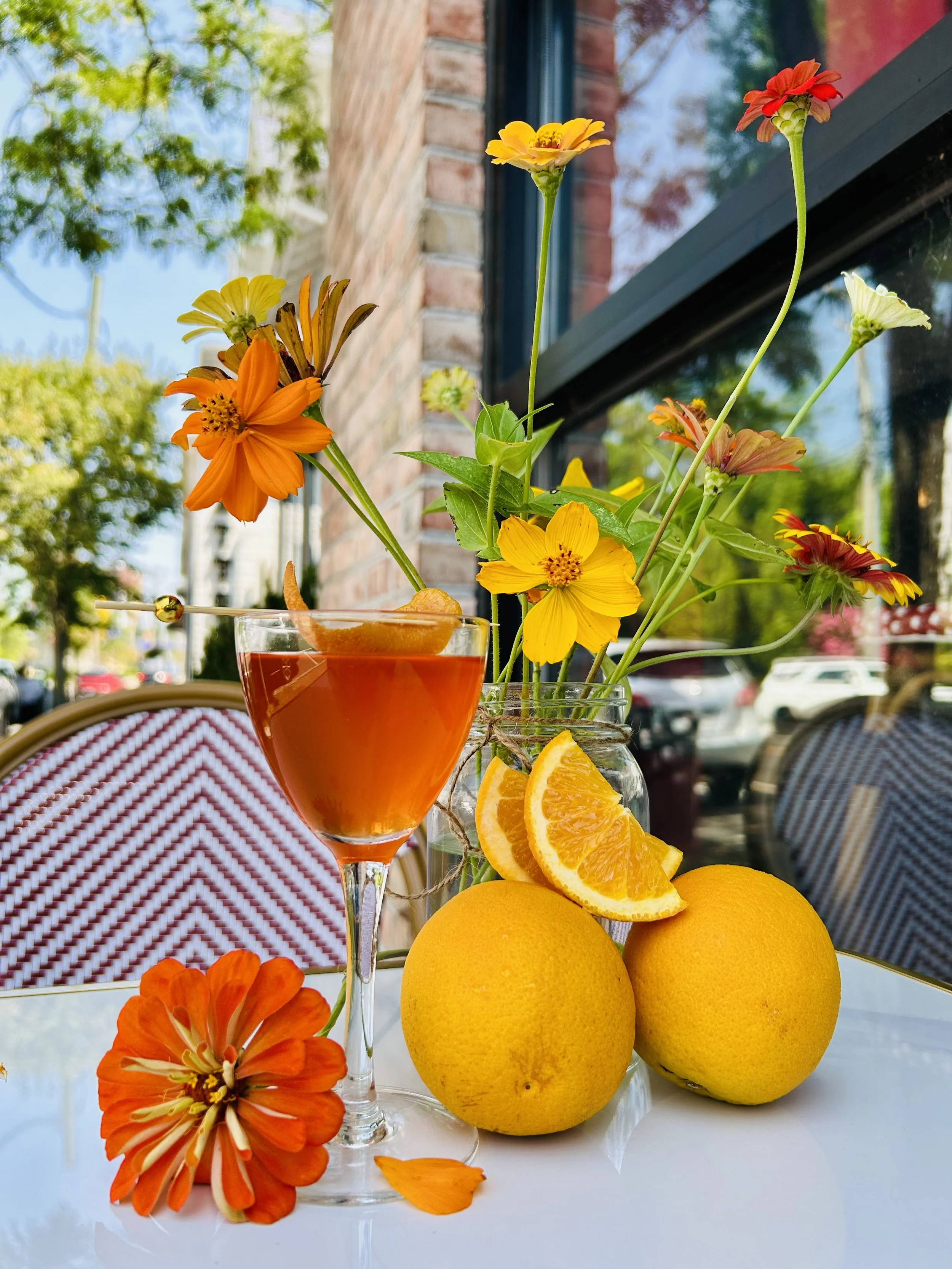 A colorful arrangement of flowers, oranges, and a glass of orange-colored drink on a white table outdoors.