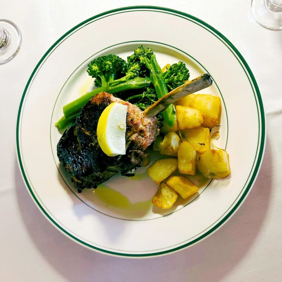 Grilled lamb chop with lemon wedge, served with broccoli and fried potatoes on a white plate with green rim
