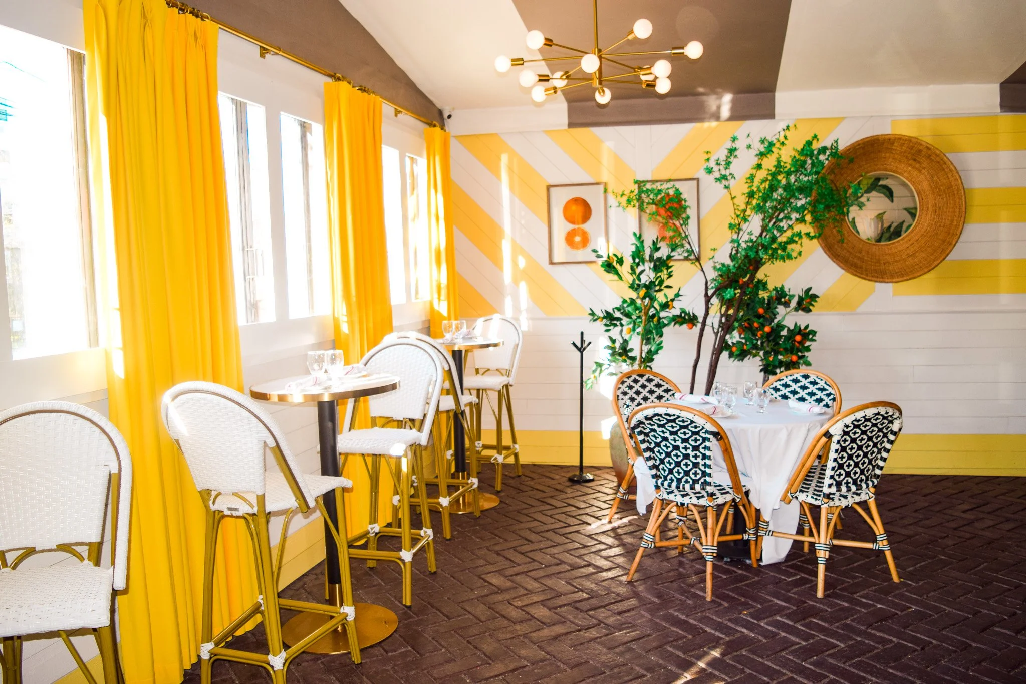 Brightly decorated dining room with yellow curtains, white chairs, and a round table set for dining. There is a wall with a yellow and white diagonal stripe pattern, two framed pictures, a large round mirror, and lush green plants. A modern chandelie