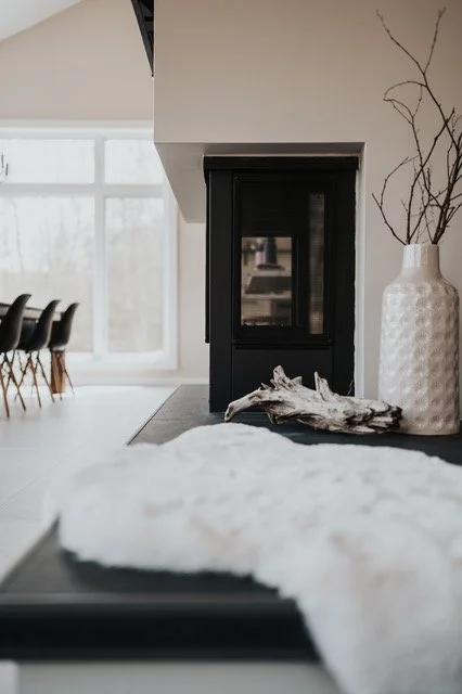 Intérieur moderne avec cheminée noire, grande vase blanche avec branches sèches, pièce lumineuse avec fenêtres et mobilier en arrière-plan.