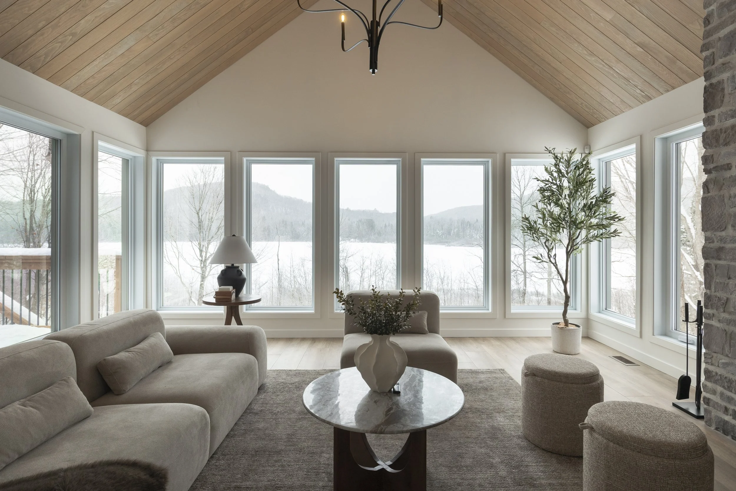 Salon avec vue sur une vallée enneigée, canapé beige, table basse en marbre avec vase, fauteuils et plantes, plafond en bois