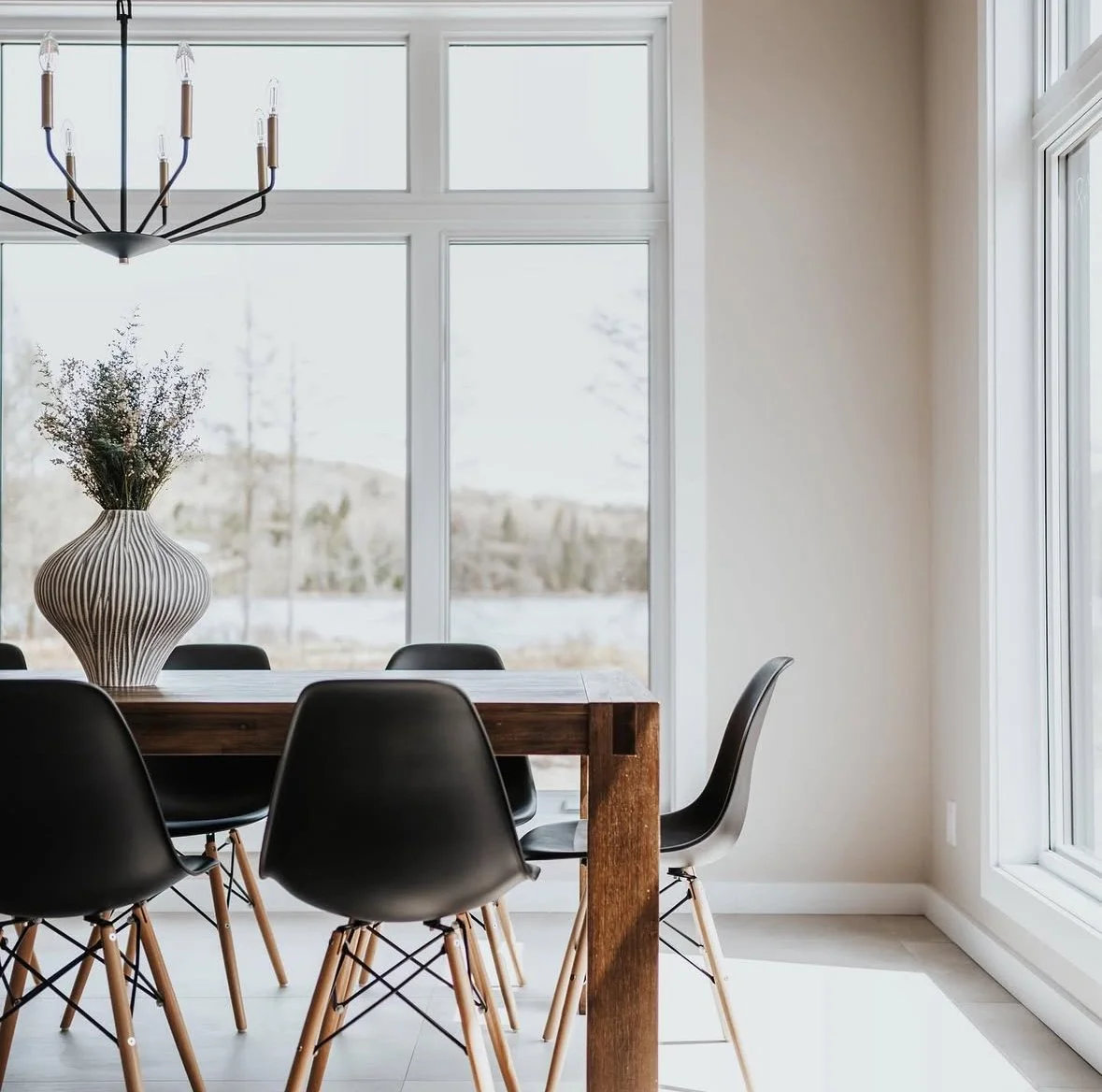 Salle à manger avec un grand vase en céramique contenant des fleurs, table en bois avec des chaises noires, grande baie vitrée laissant entrer beaucoup de lumière naturelle.