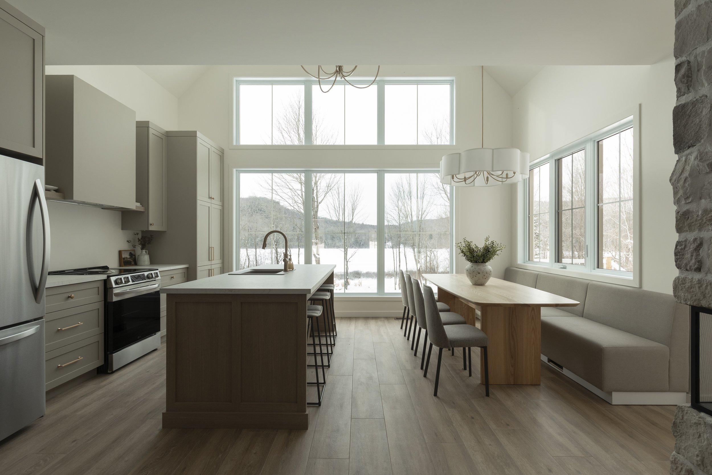 Cuisine moderne avec grande baie vitrée donnant sur un paysage hivernal, comptoir central avec évier, table en bois avec chaises grises, banc intégré, décor épuré et lumineux.