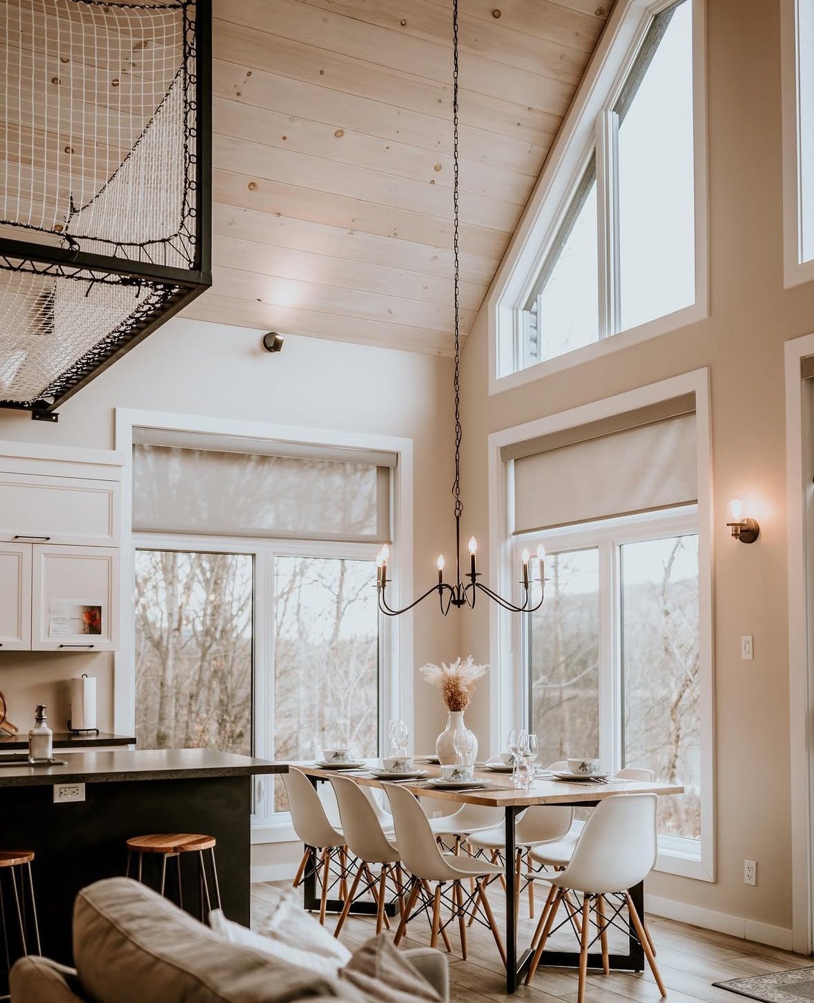 Salle à manger avec grande baie vitrée, chandelier suspendu et décor minimaliste, vu depuis un salon avec canapé beige.