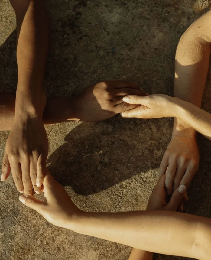 Four hands holding each other's fingers in a circle on a sunlit ground.