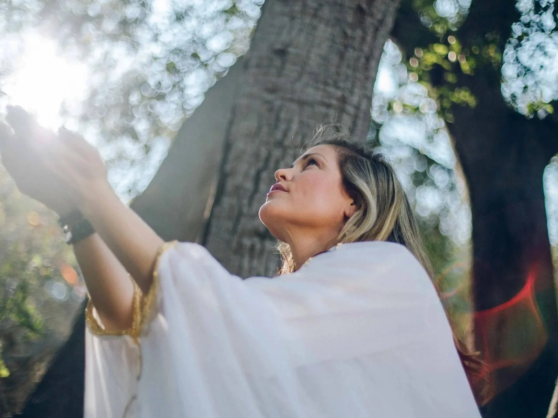 A woman with long, blonde hair looking upward while standing near a large tree in a forested area during daylight.
