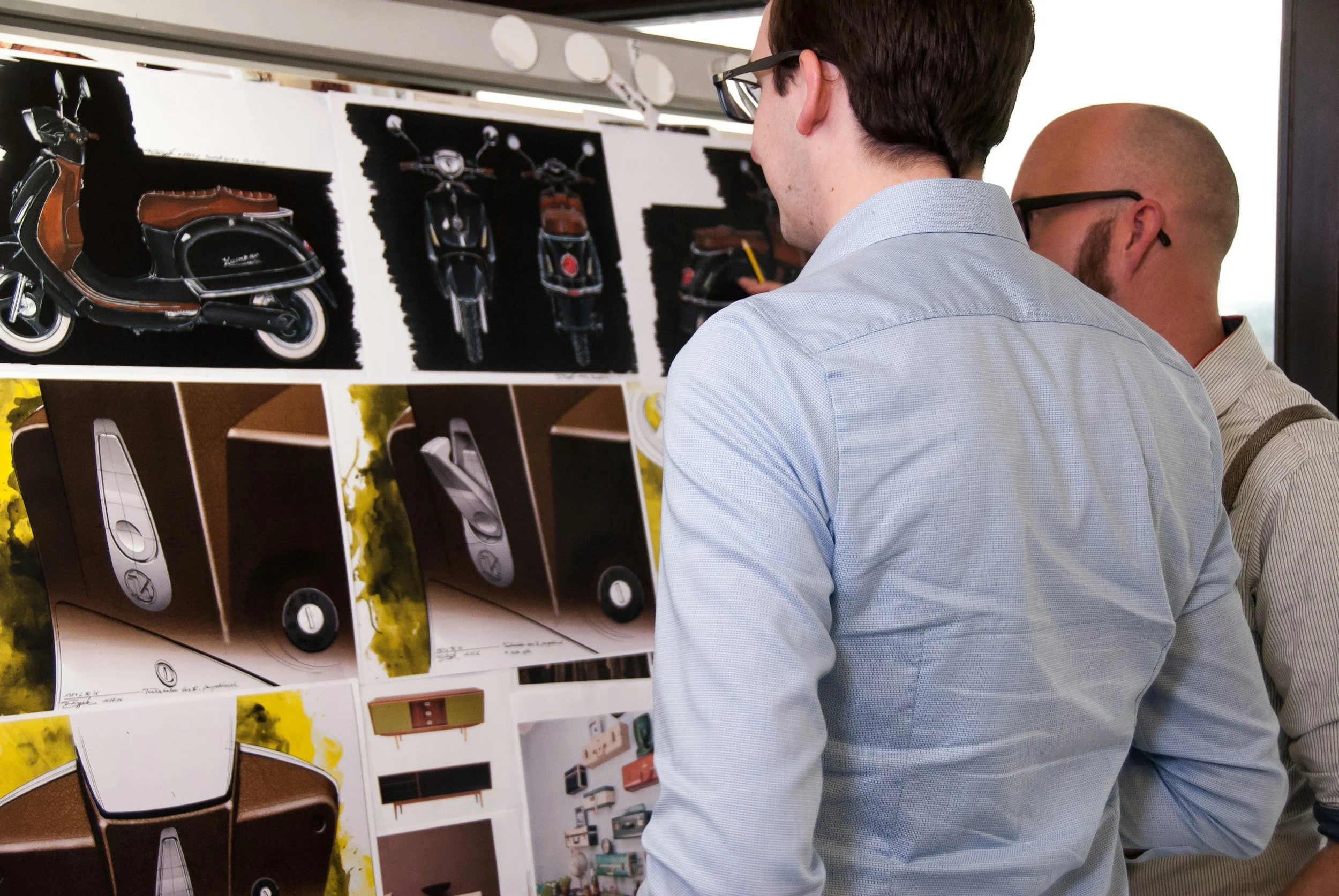 Two men looking at a display board with pictures of vintage scooters and furniture.