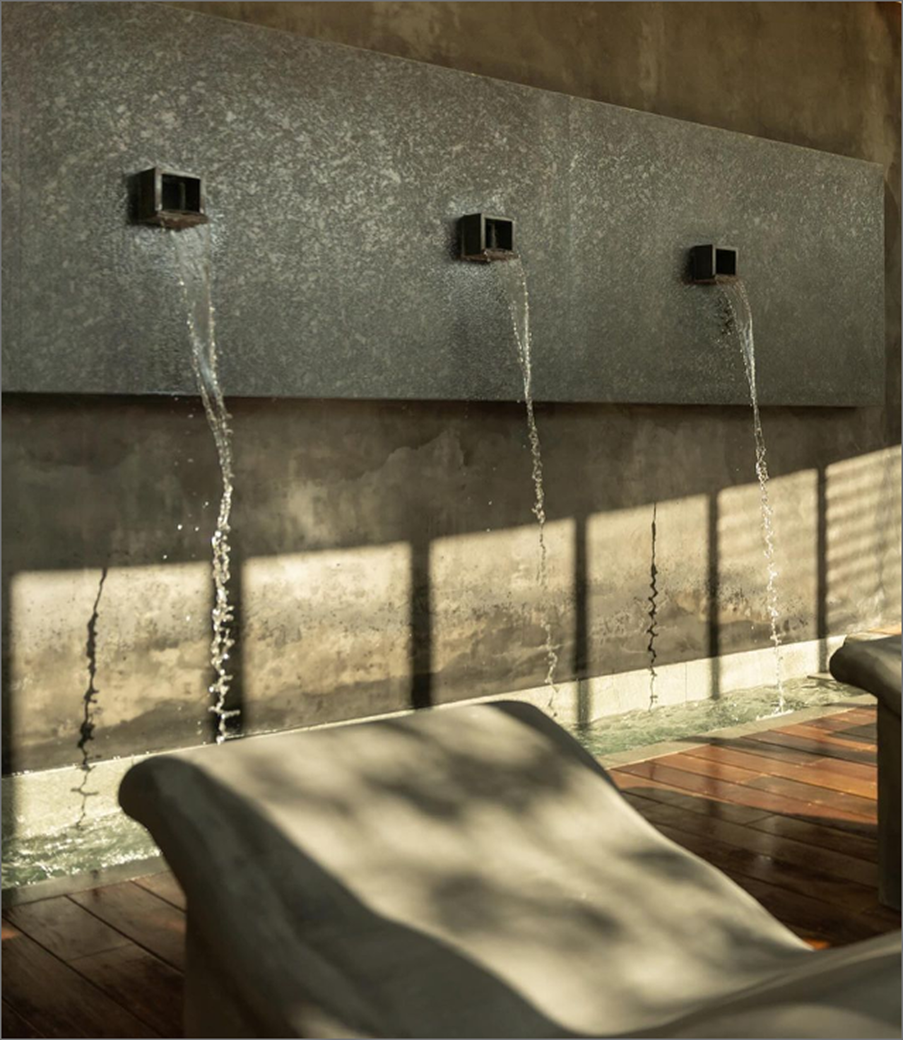 Three water spouts mounted on a textured gray wall, with water flowing out of each, casting shadows on the wall below, and benches in the foreground.