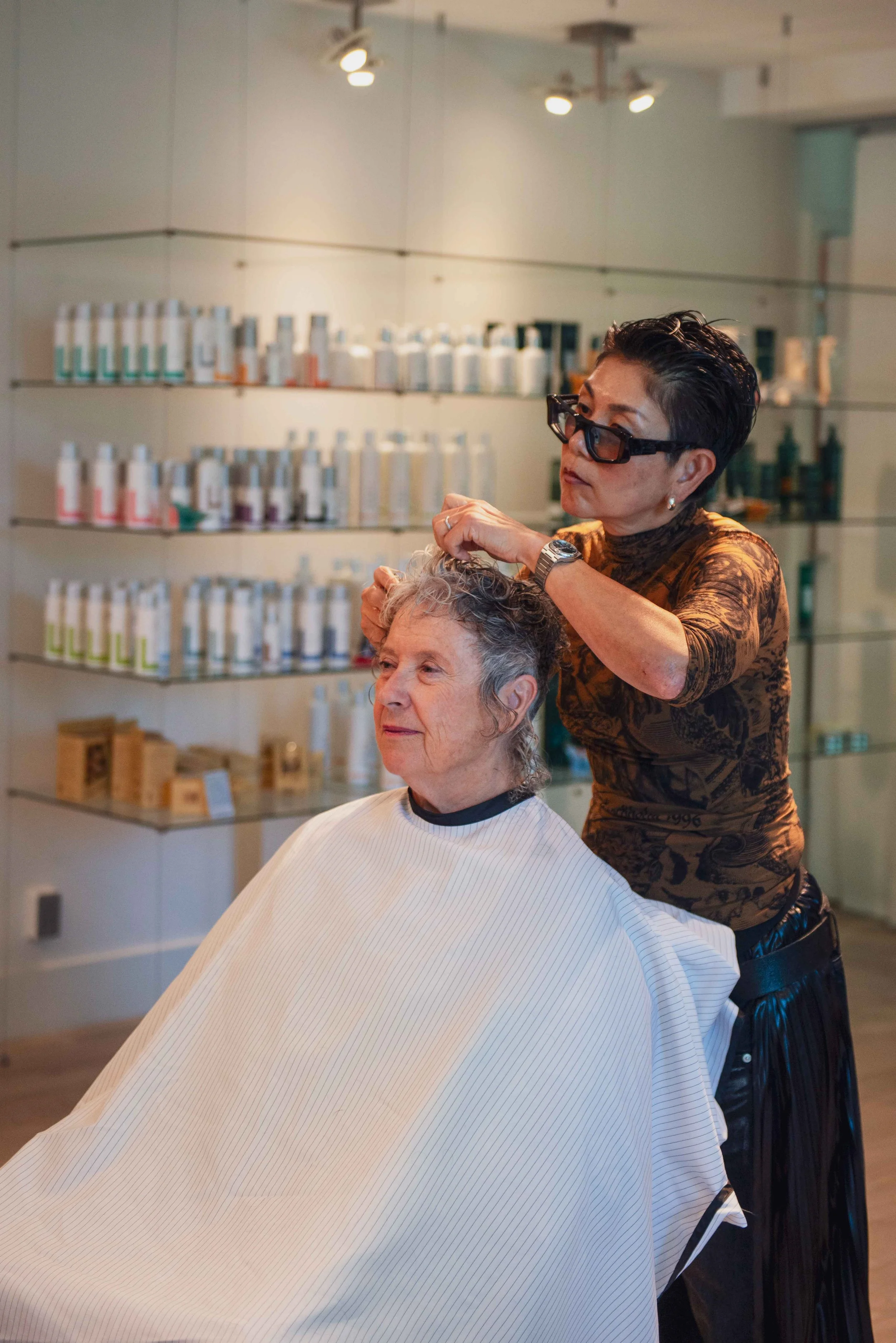 A hairdresser cutting an elderly woman's hair in a salon, with shelves of hair products in the background.