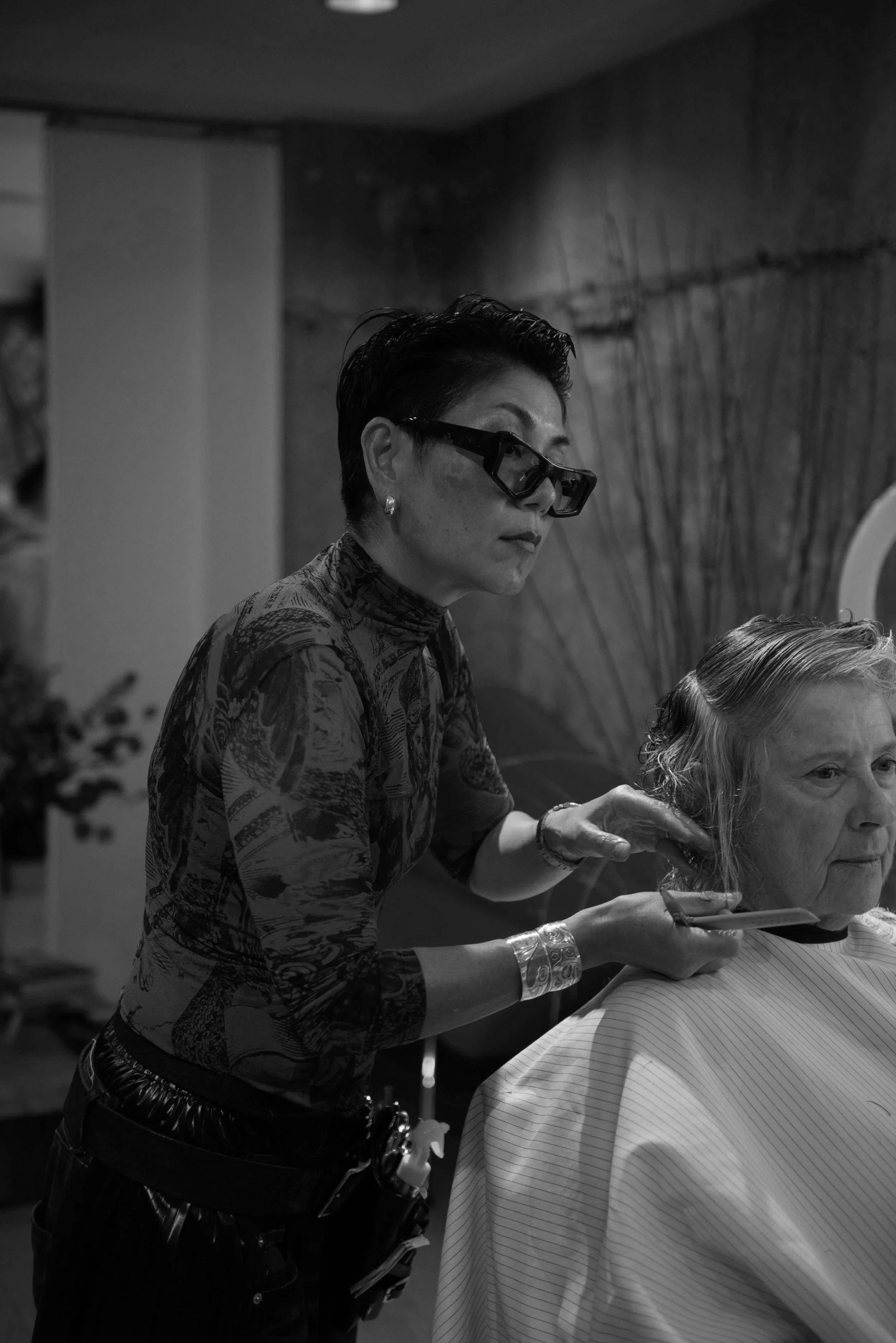 A hairstylist, wearing glasses and patterned top, cuts an elderly woman's hair in a salon.