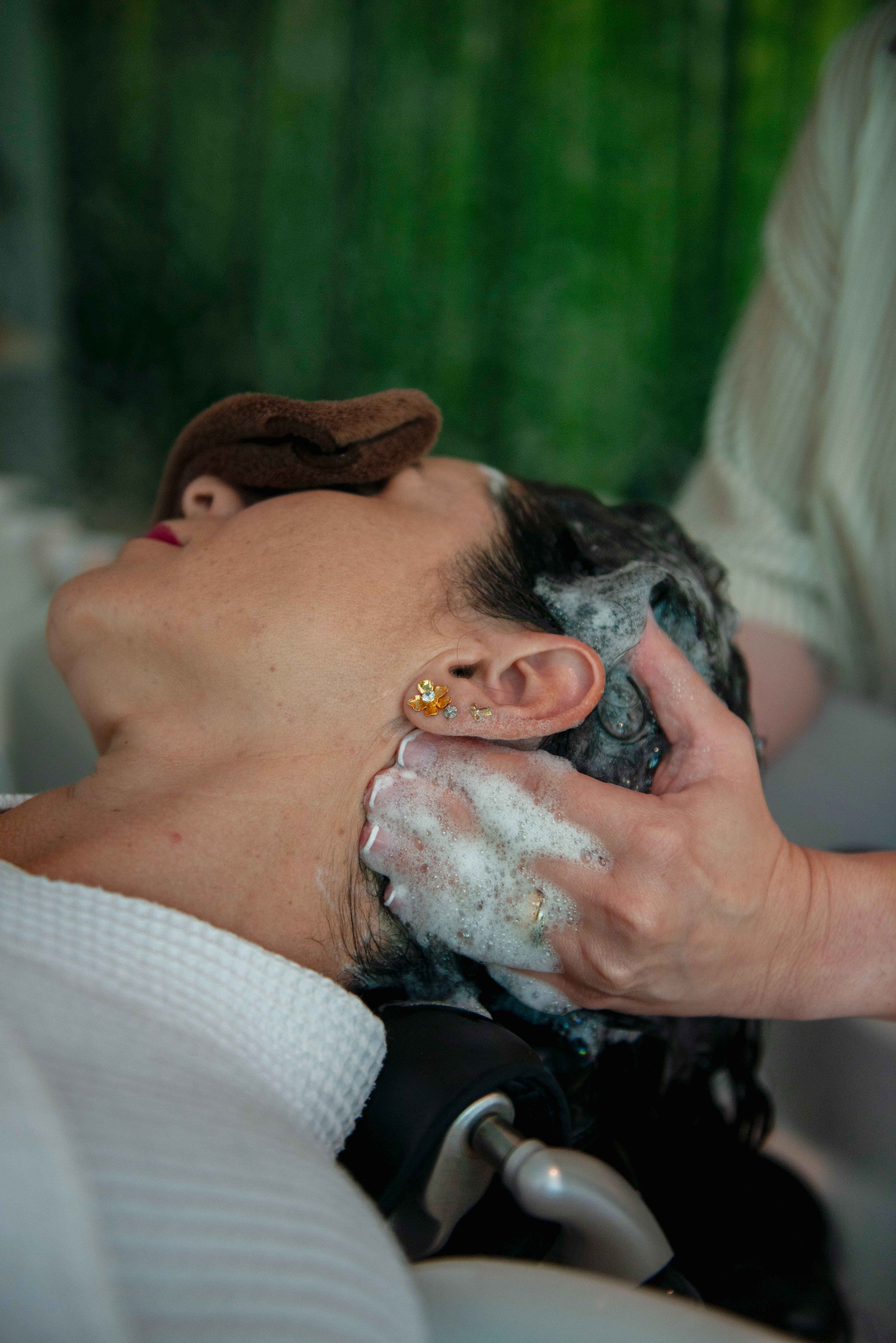 Woman with dark hair getting a head massage in a spa, with a towel on her forehead, earrings, and bubbles in her hair.