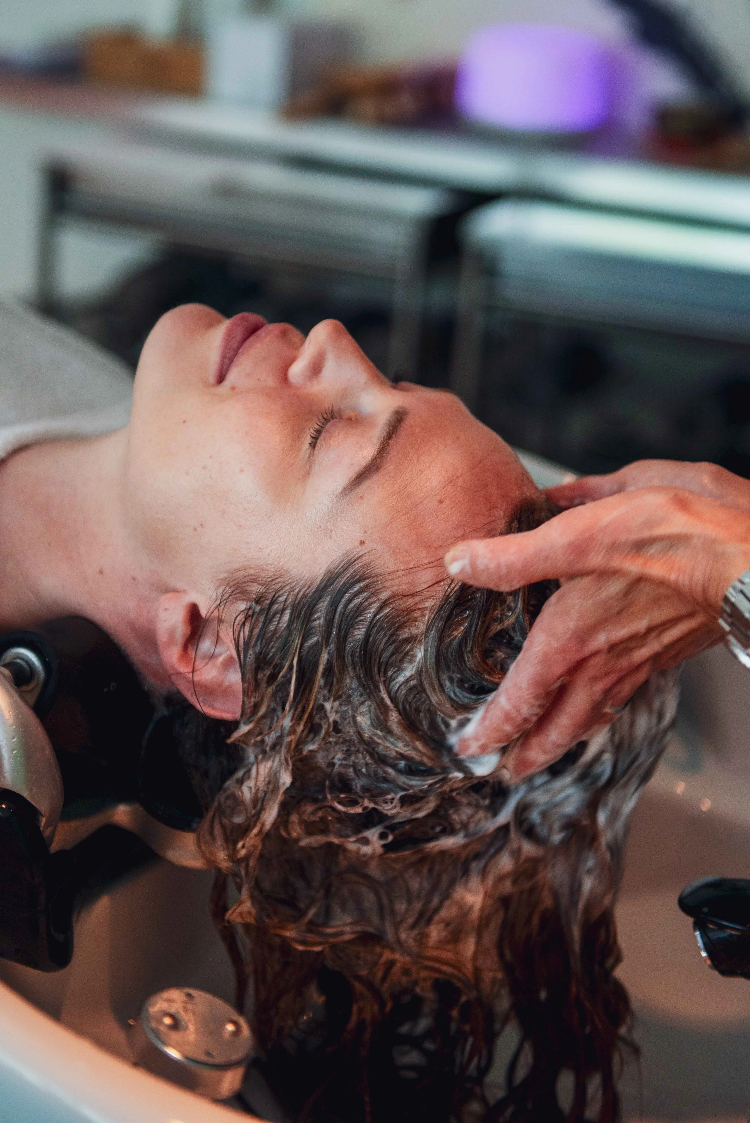 Person receiving a hair wash at a salon, leaning back with eyes closed as a stylist shampoos their curly hair.