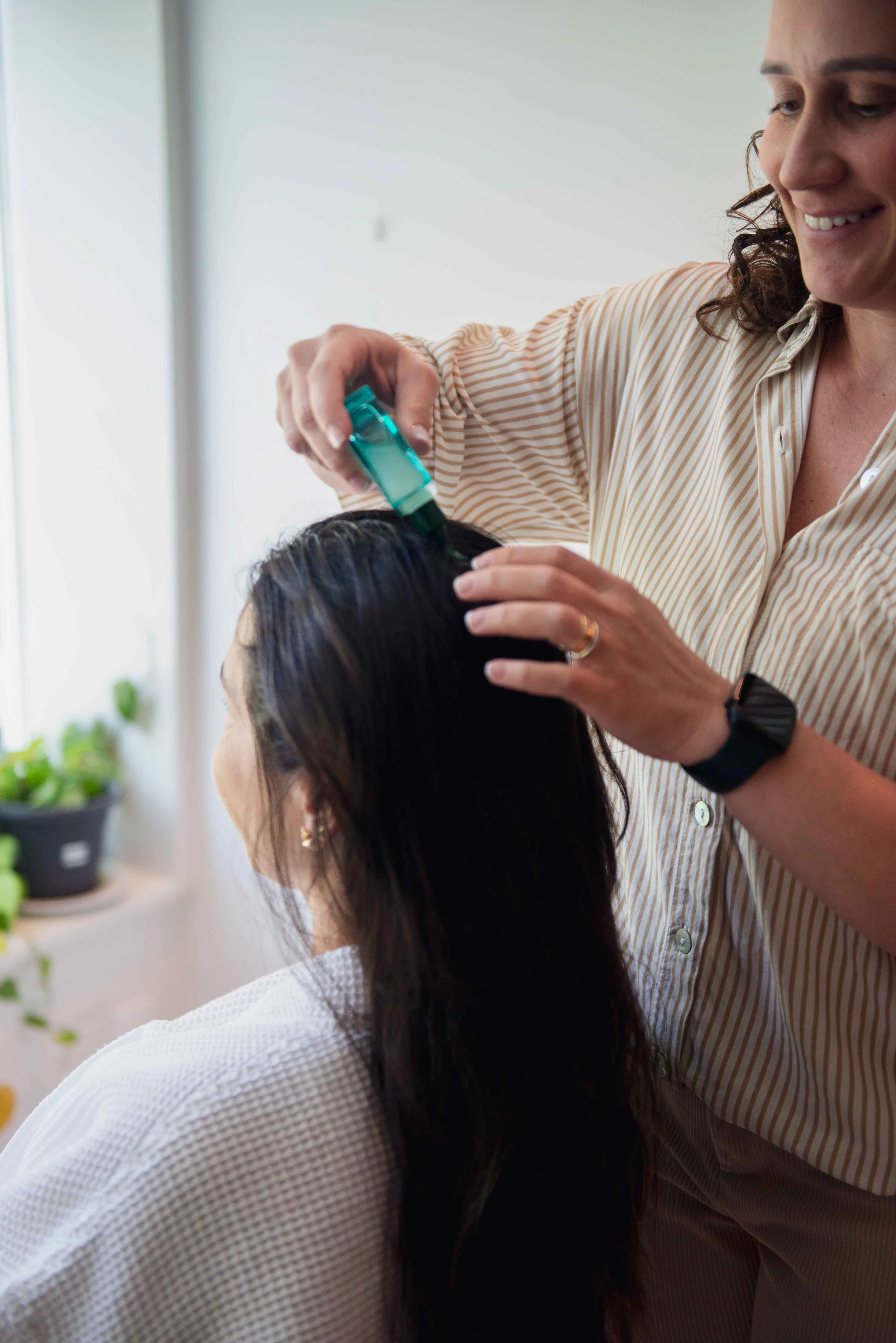 Woman applying hair treatment to a woman's hair in a well-lit room with plants in the background.
