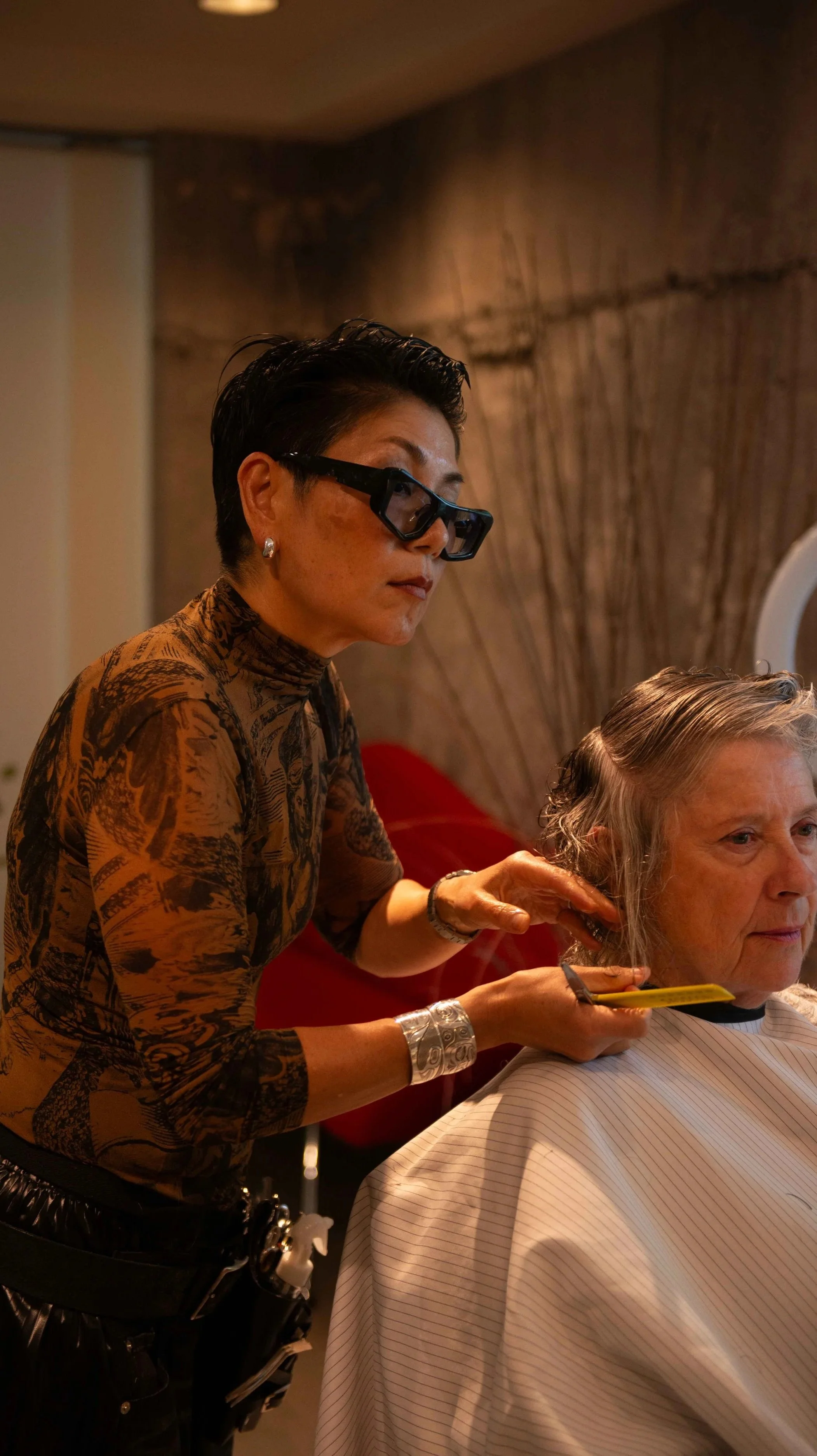 A woman with short black hair, wearing glasses, is styling an older woman's gray hair in a salon.