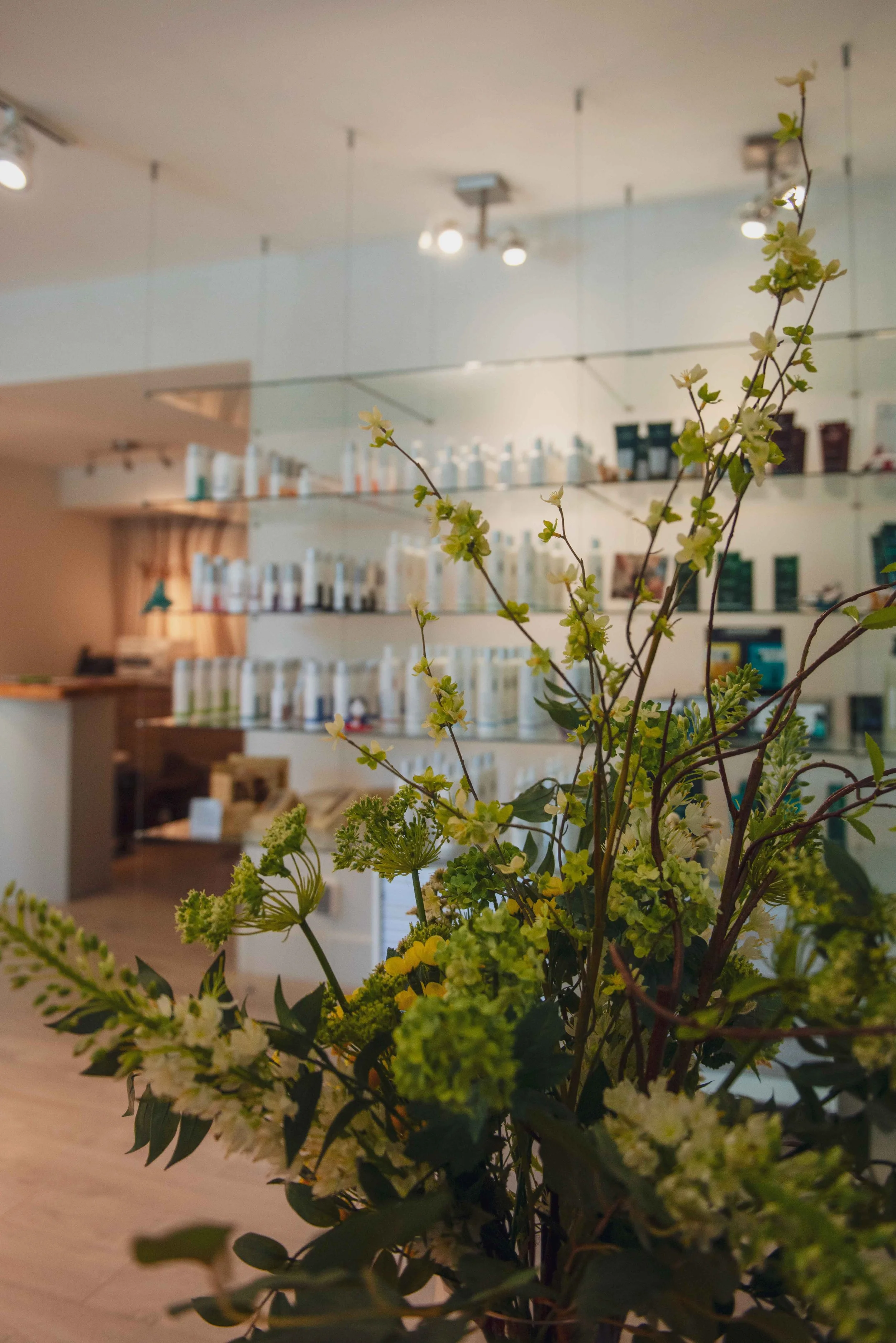 Close-up of a floral arrangement with green and white flowers in the foreground. The background shows a room with shelves lined with bottles and products, and track lighting on the ceiling.